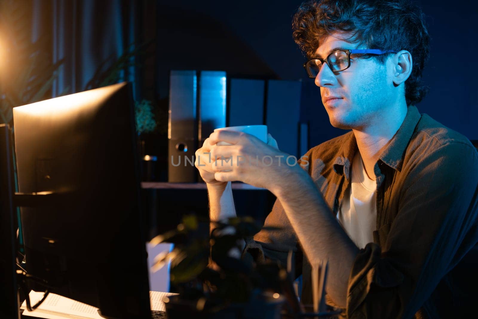 Young businessman working creative startup project job, drinking hot drink with coffee or tea while focusing on pc at neon light modern office. Thinking marketing plan online at night time. Gusher.