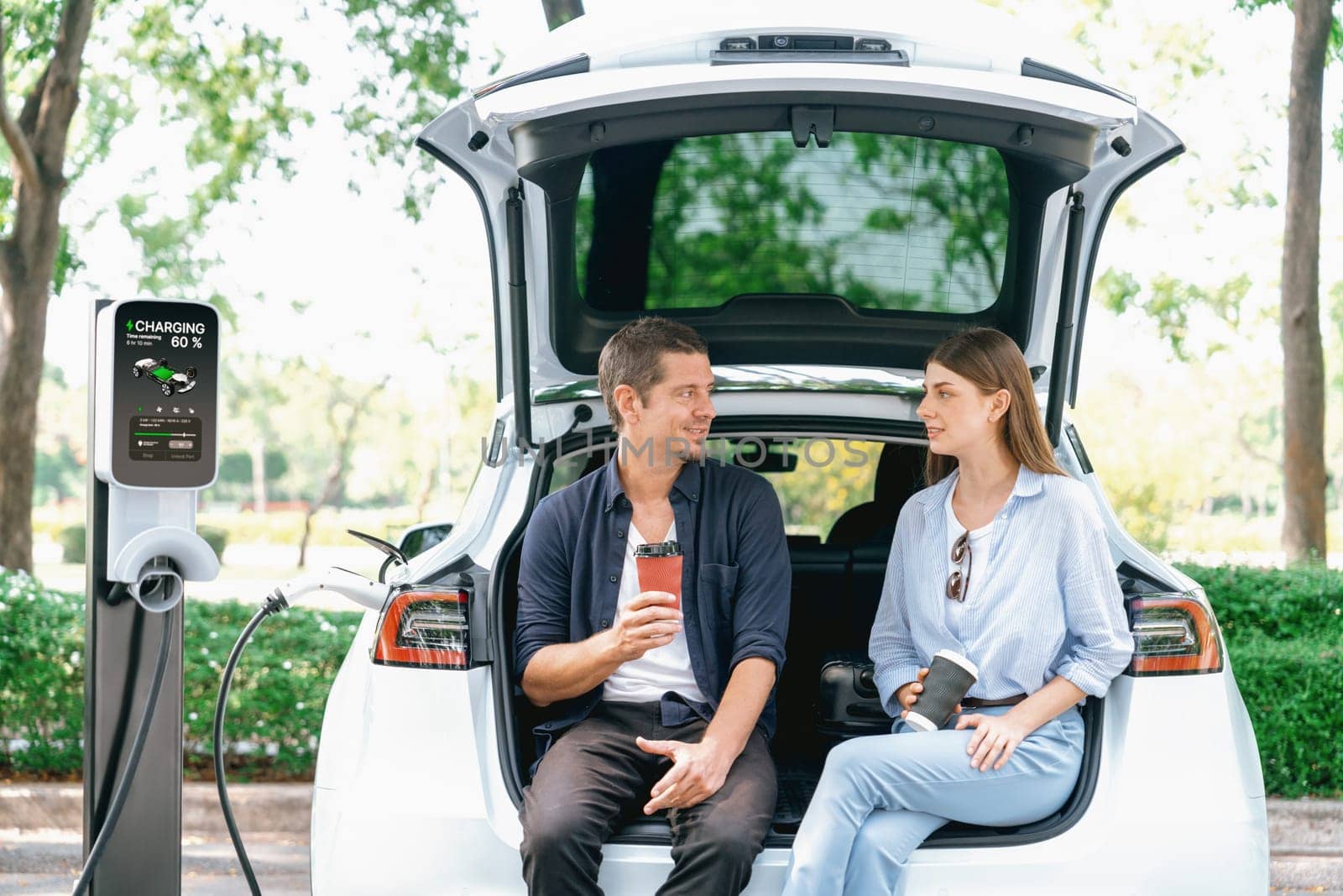 Lovely young couple drinking coffee while recharging battery for electric car during road trip travel EV car in natural forest or national park. Eco friendly travel during vacation and holiday. Exalt