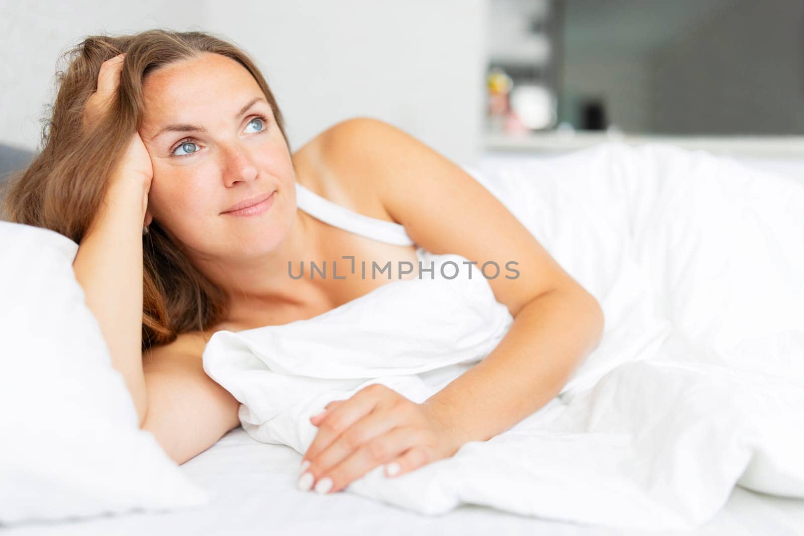 A thoughtful woman lies in bed with a serene expression, wrapped in white linens, enjoying a peaceful moment.