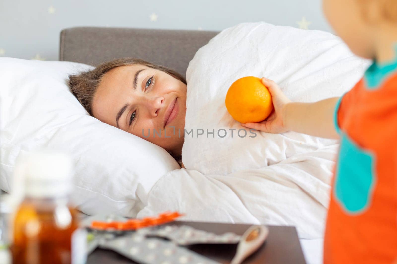 A caring young child hands an orange to their ill mother lying in bed, with medication visible on the bedside.