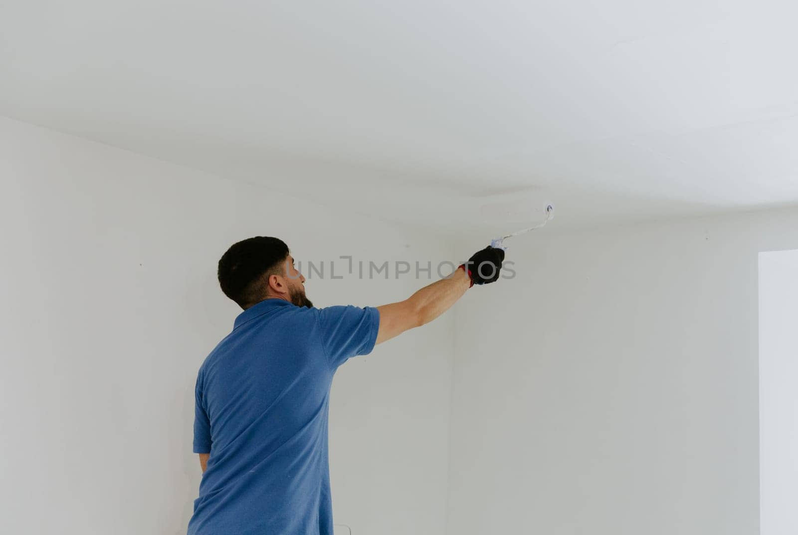 One young handsome brown-haired Caucasian man in a blue T-shirt stands from the back and paints the ceiling next to the doorway with a roller of white paint, close-up side view with selective focus and copy space on the right.