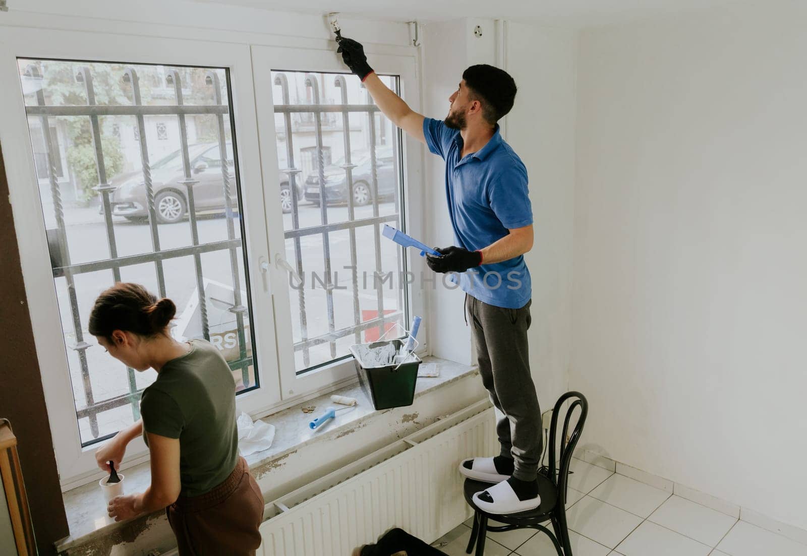 A young handsome couple of a dark-haired bearded Caucasian man and a girl stands sideways by the window and paints the window opening with a brush with white paint, close-up side view with selective focus.