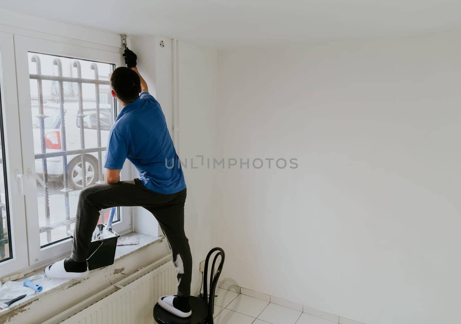 One young handsome dark-haired bearded Caucasian man in a blue T-shirt stands on a chair sideways by the window and paints the window opening with a brush with white paint, close-up side view with selective focus.