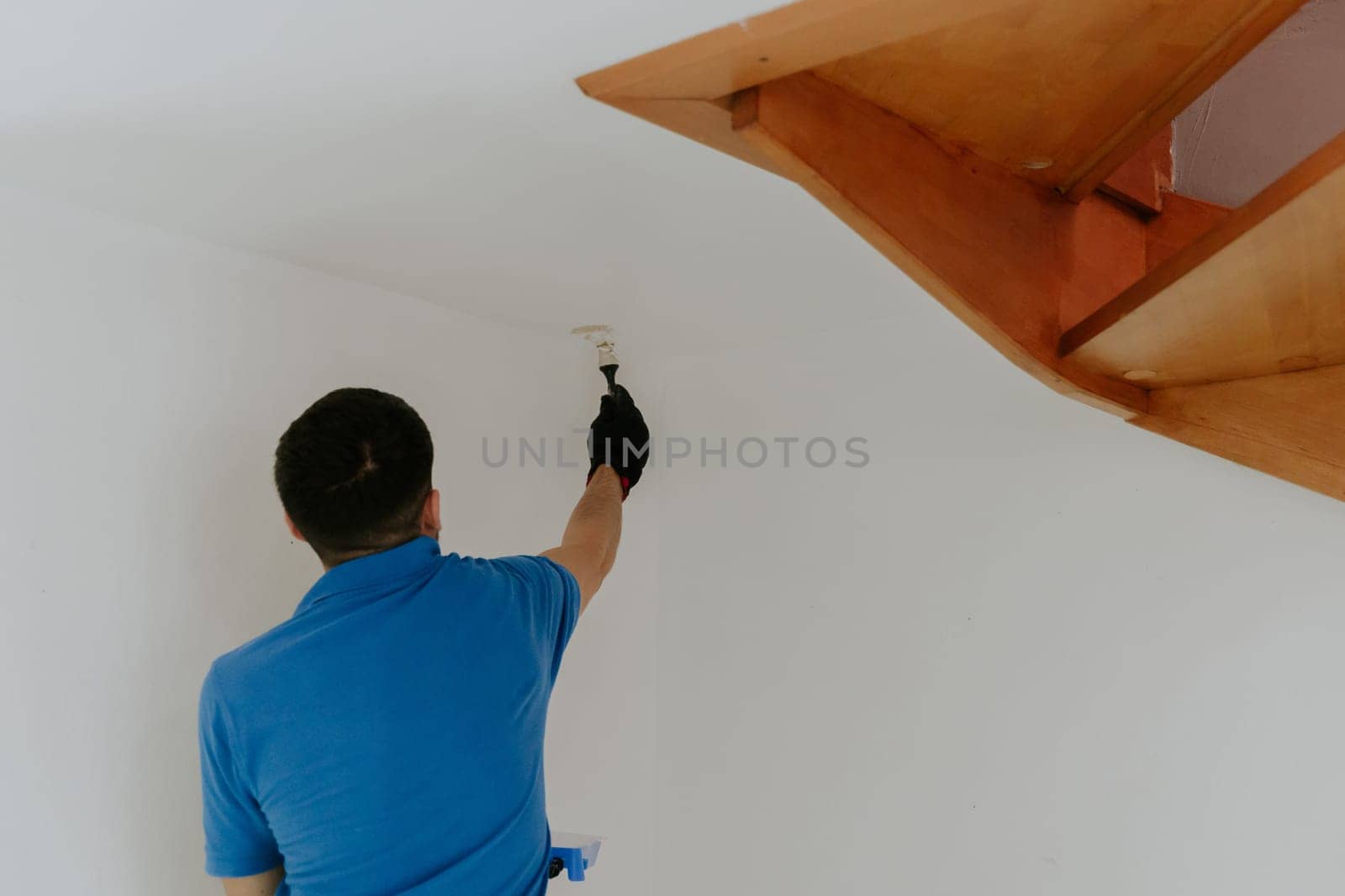 One young handsome dark-haired Caucasian man in a blue T-shirt stands from the back and paints with a brush with white paint the corners of the wall under the wooden stairs, close-up side view with selective focus.