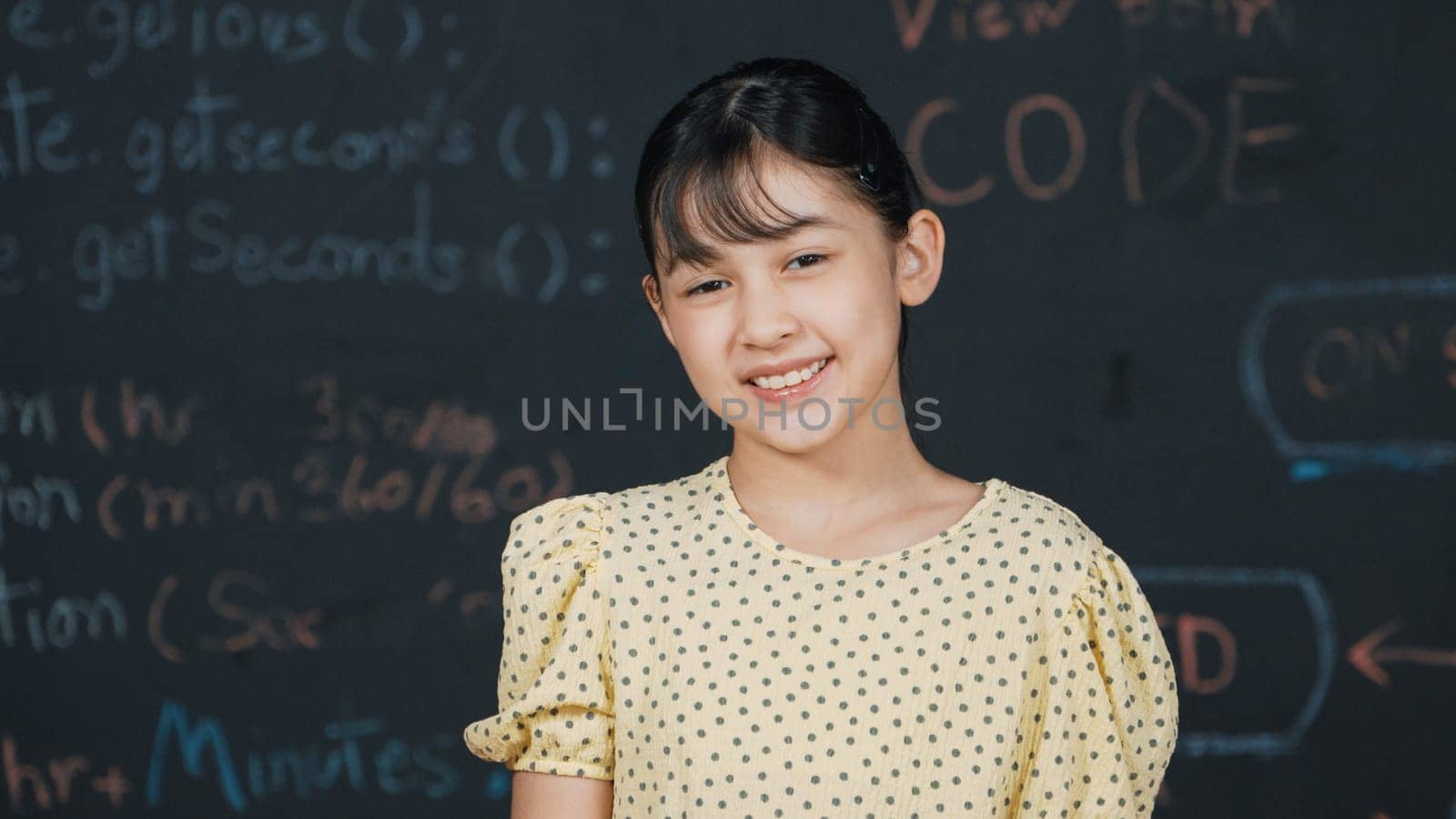 Cute happy girl smiling to camera while standing at blackboard with engineering prompt or coding, programing system written in STEM technology classroom. Smart student looking at camera. Edification.