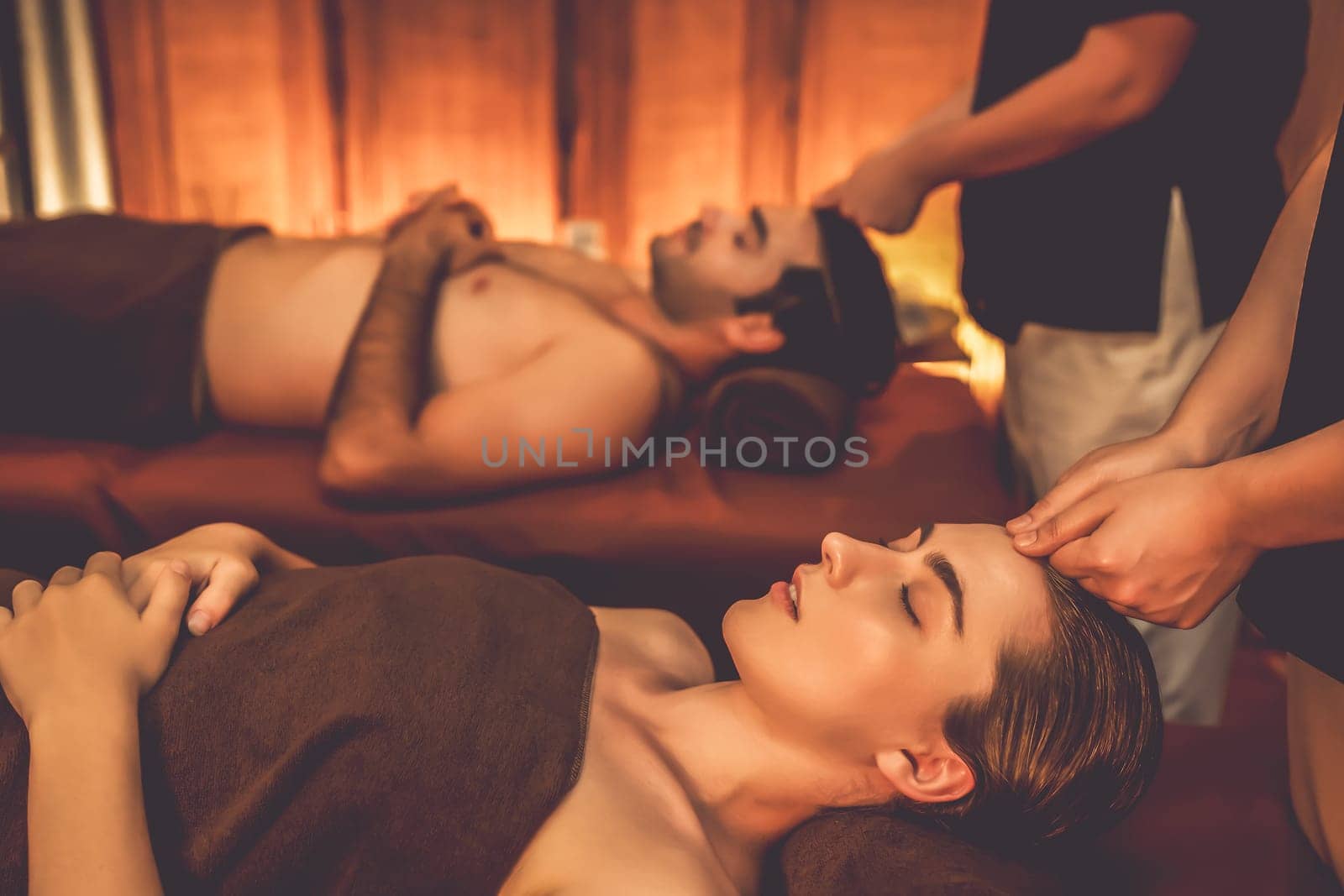 Couple customer enjoying relaxing anti-stress head massage and pampering facial beauty skin recreation leisure in warm candle lighting ambient salon spa in luxury resort or hotel. Quiescent