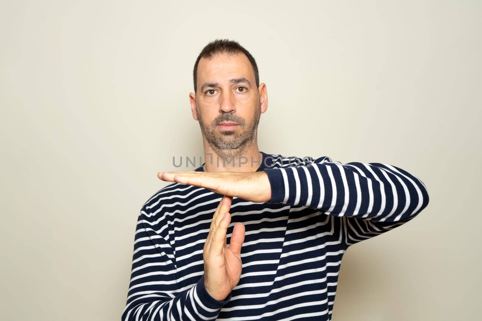 Bearded Hispanic man in his 40s wearing a striped sweater calling for a time out with his hands isolated on a beige background, he needs a break from the fast pace of his life. by Barriolo82