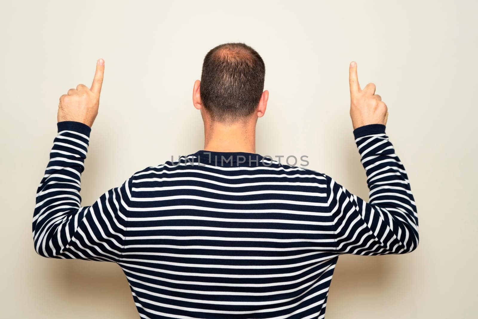 Hispanic man wearing a striped sweater pointing up with index fingers with his back to the camera, isolated on beige background.