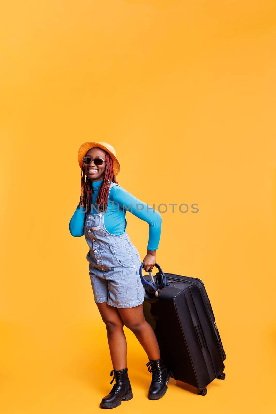 Young adult with sunglasses and hat posing on camera, carrying trolley bags to travel on holiday trip. Happy girl feeling excited about leaving on vacation, wearing modern glasses.