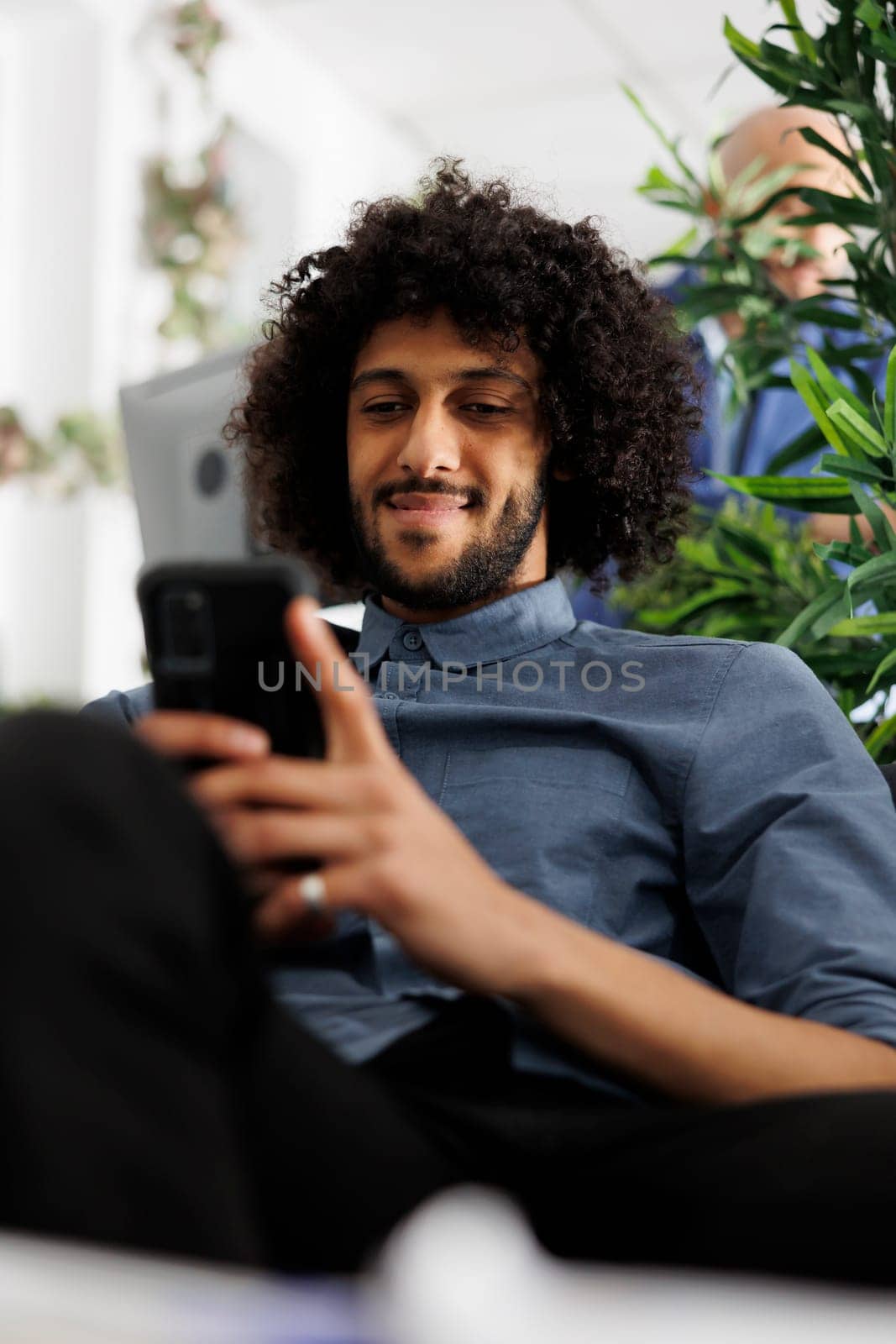 Smiling arab executive manager texting in social network on smartphone in business office. Start up company employee chatting on mobile phone while sitting on couch in coworking space