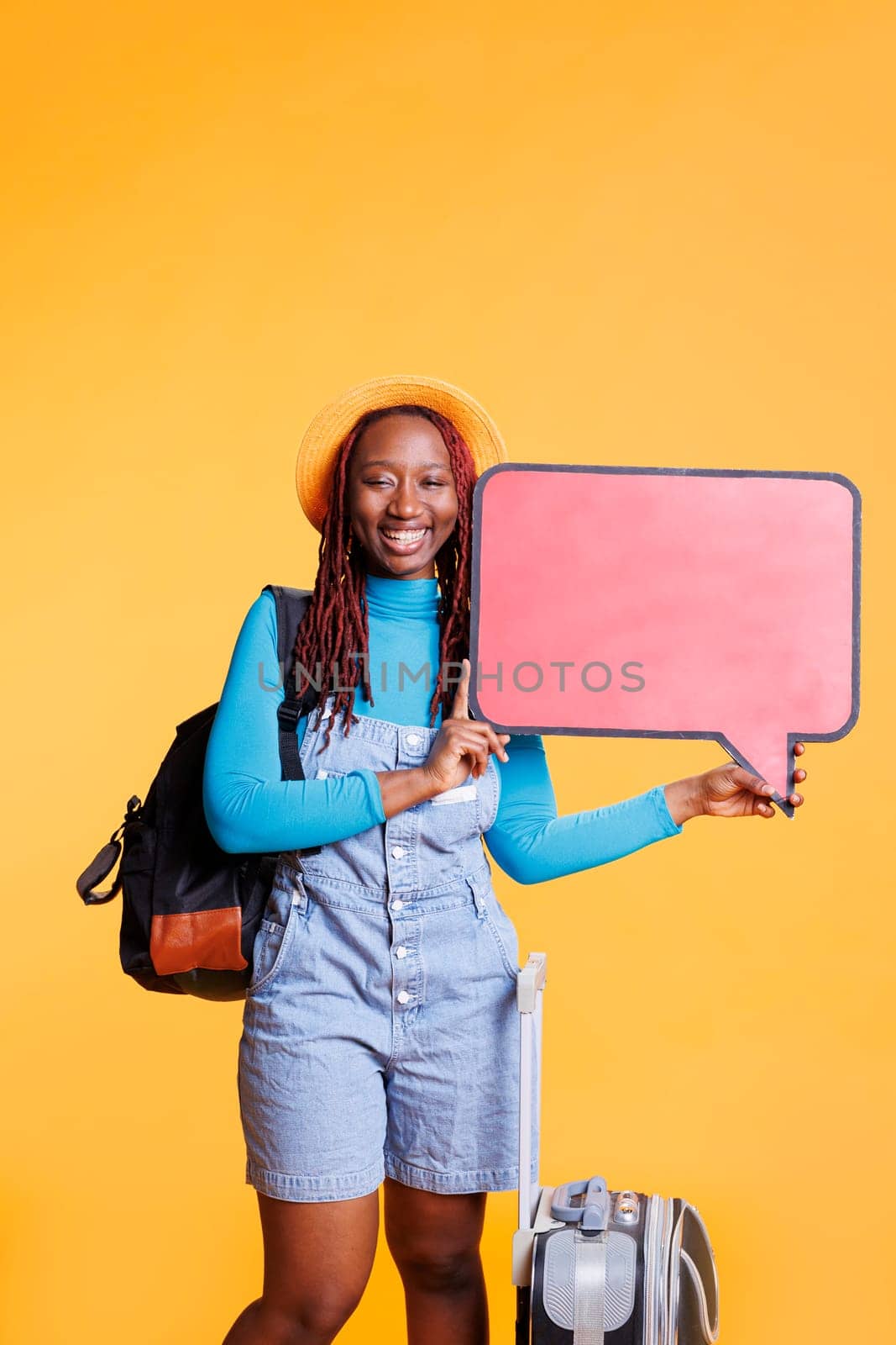 Happy tourist with speech bubble icon creating ad, travelling on holiday trip with bags. Young female model showing copyspace empty cardboard with advertisement in studio, isolated board.