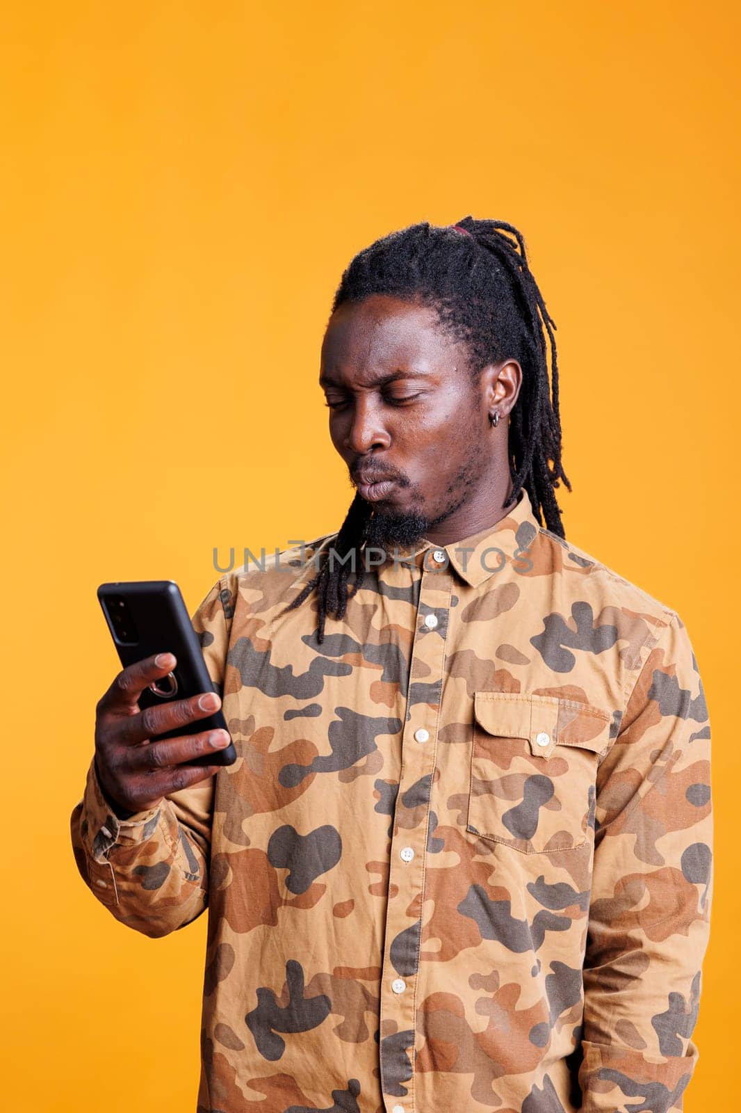 African american man reading good news on smartphone, scrolling through social media in studio over yellow background. Cheerful person searching information on phone, browsing on internet