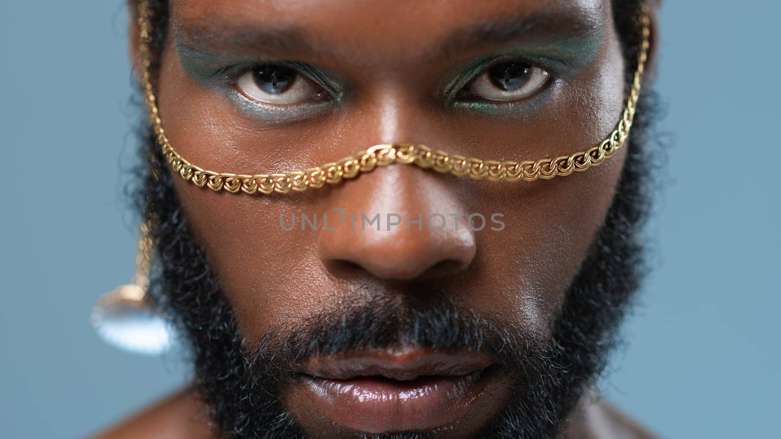 Closeup portrait of bearded African American gay man with blue eyeshadow and golden accessory on face. Serious homosexual adult male with makeup looking at camera on blue background.