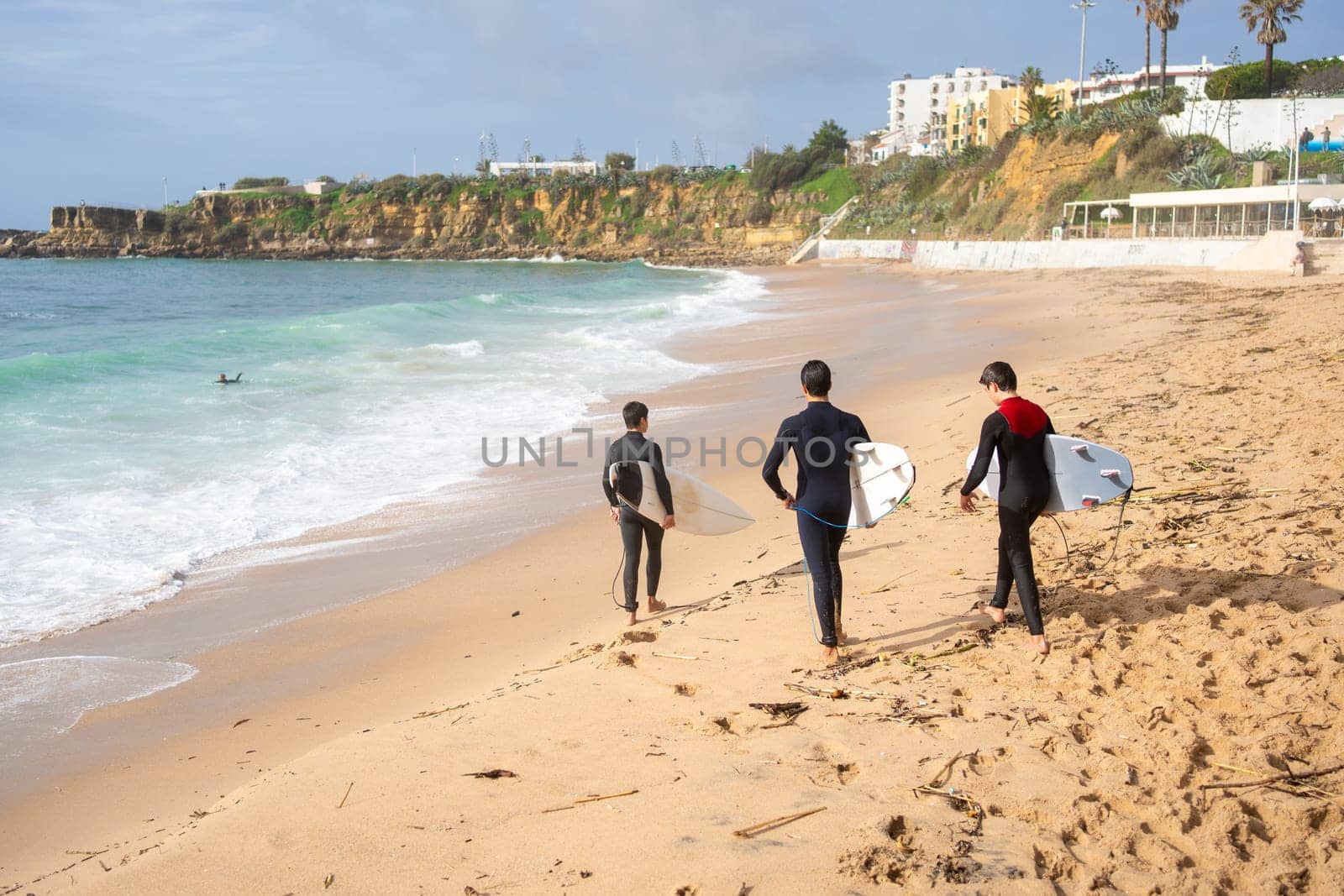 Back view of anonymous surfers walking with surfboards along sea waves. Full body of active friends in swimwear walking on sandy beach. Summer vacation and water sport concepts.