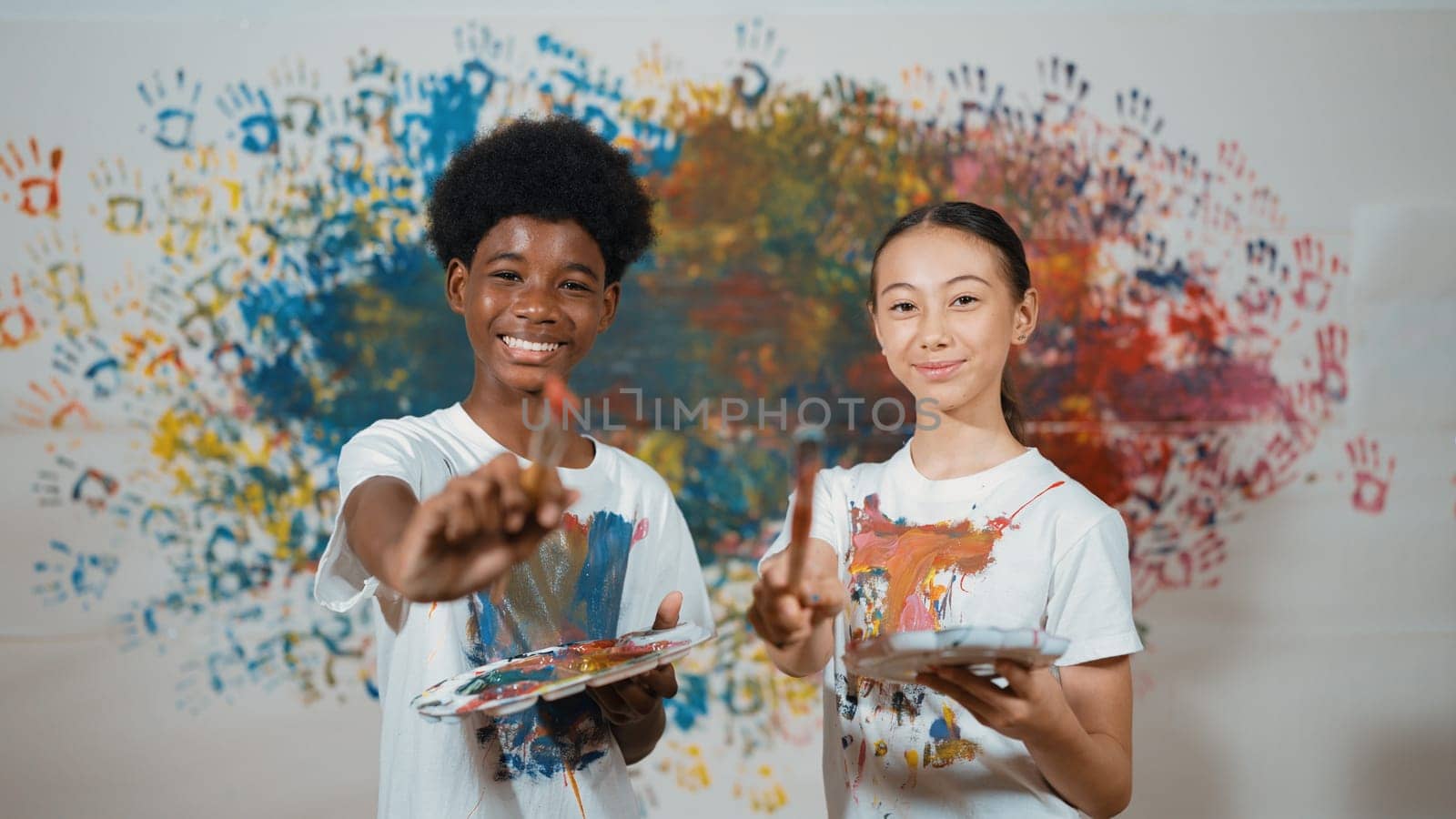 Highschool student with mixed races standing and looking at camera at art lesson. Diverse children hold the paintbrush while pose with confident at colorful stained wall. Creative style. Edification.