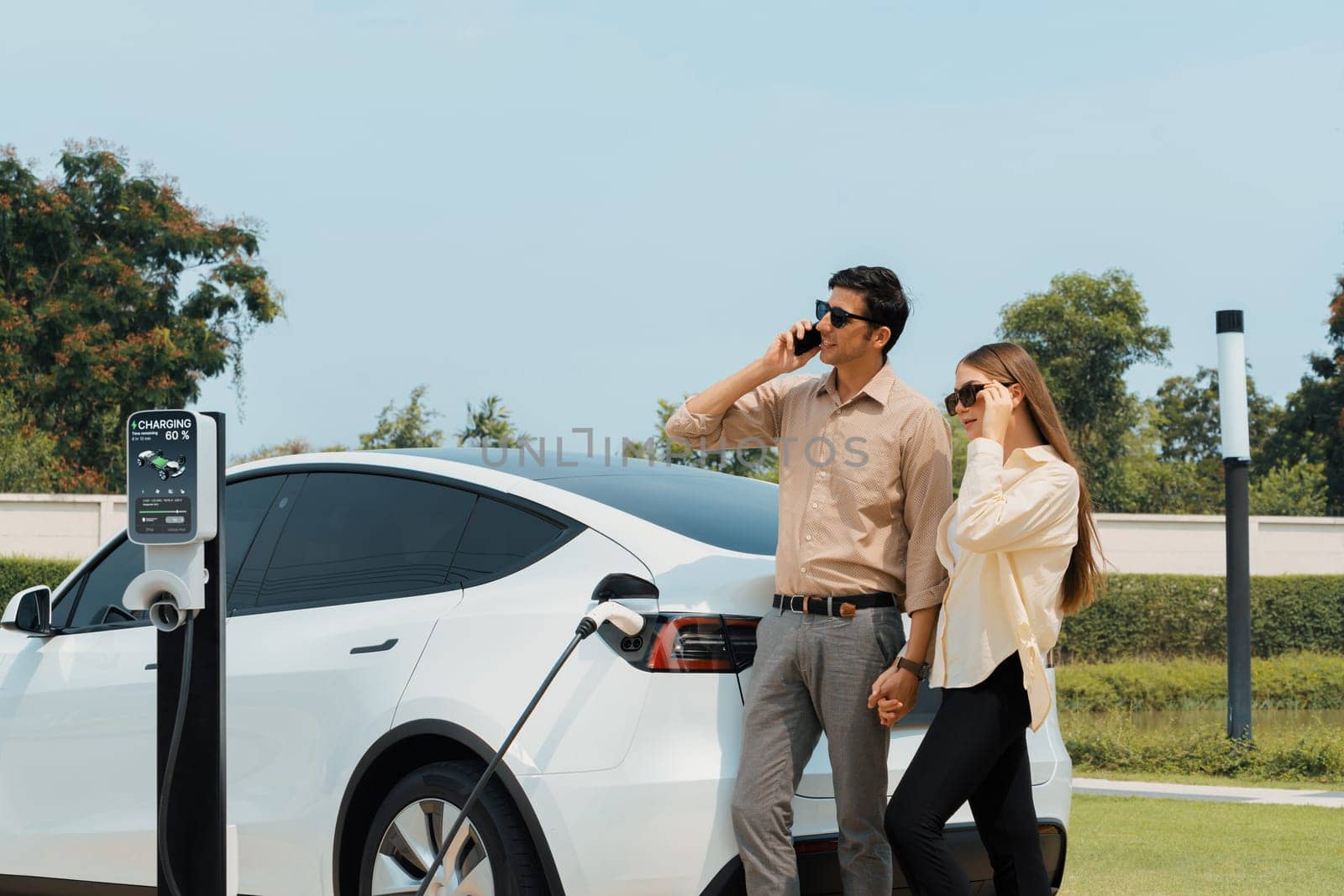 Young couple recharge her EV electric vehicle at green city park parking lot while talking on phone. Sustainable urban lifestyle for environment friendly EV car with battery charging station.Expedient