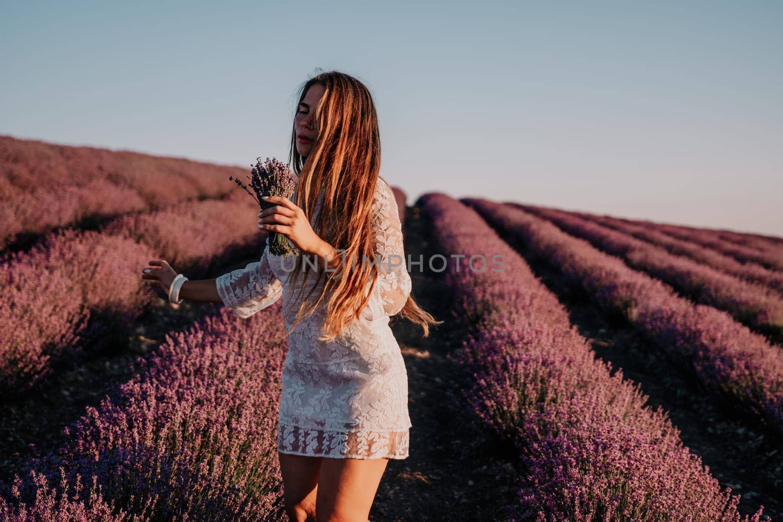 Close up portrait of young beautiful woman in a white dress and a hat is walking in the lavender field and smelling lavender bouquet.