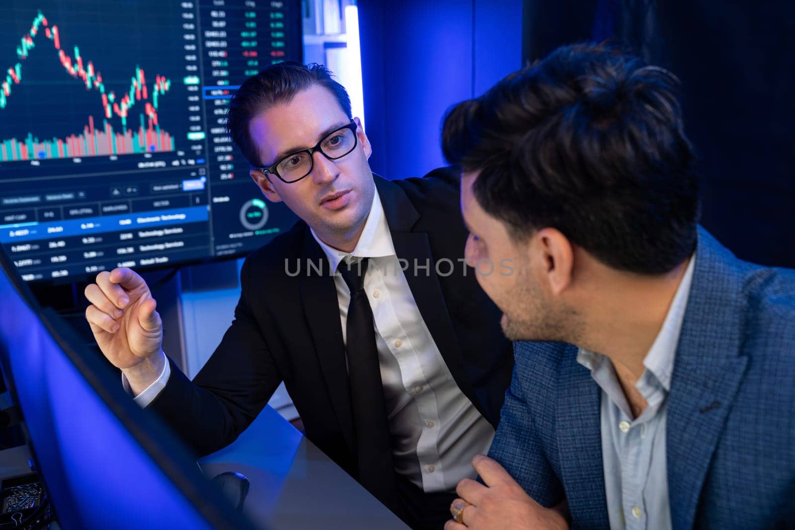 Two stock exchange traders discussing dynamic investment graph in currency rate on monitor at night. Businessman partners coffee meeting in high stock market in neon light at workplace. Sellable.