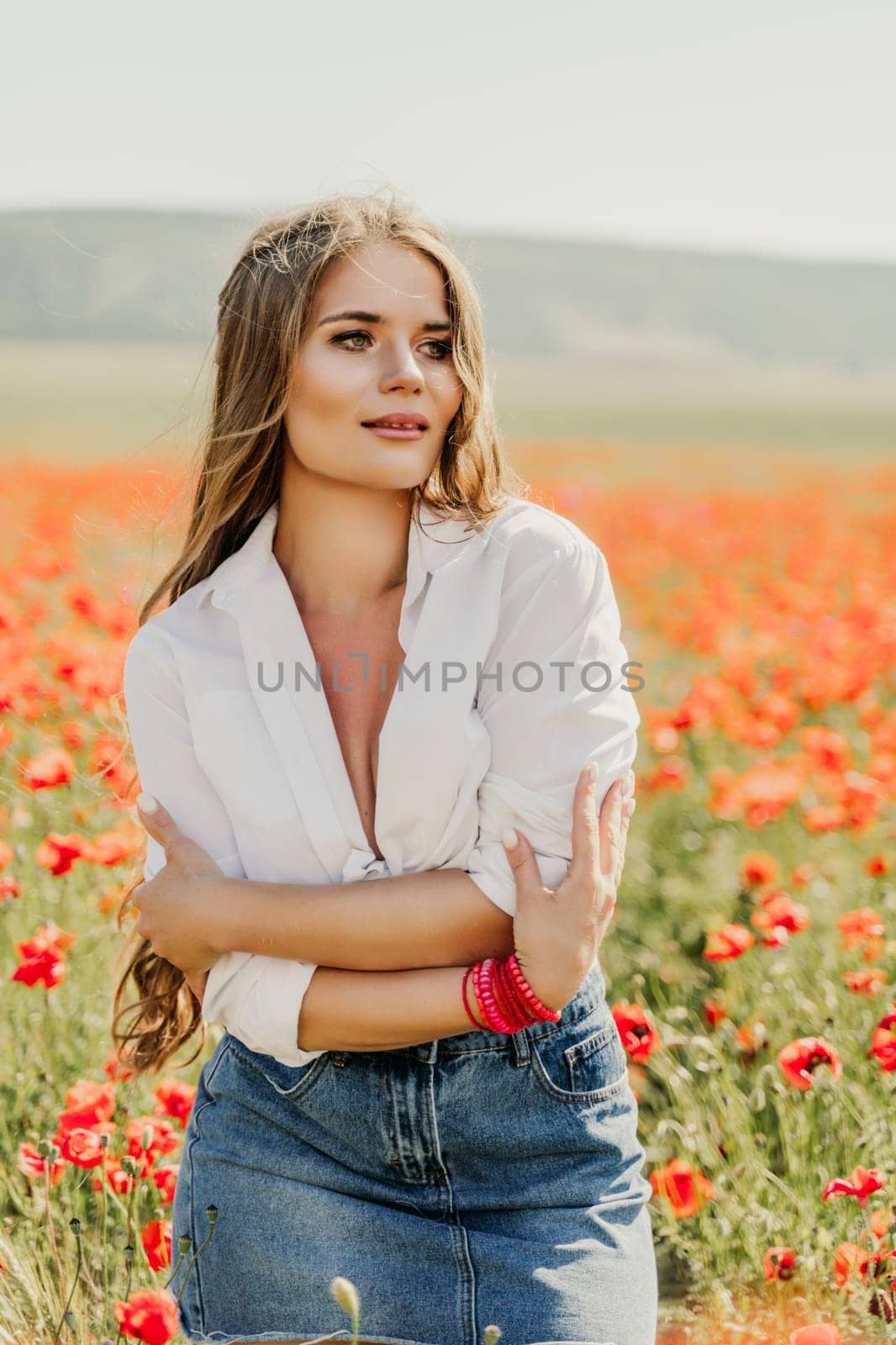 Happy woman in a poppy field in a white shirt and denim skirt with a wreath of poppies on her head posing and enjoying the poppy field. by Matiunina