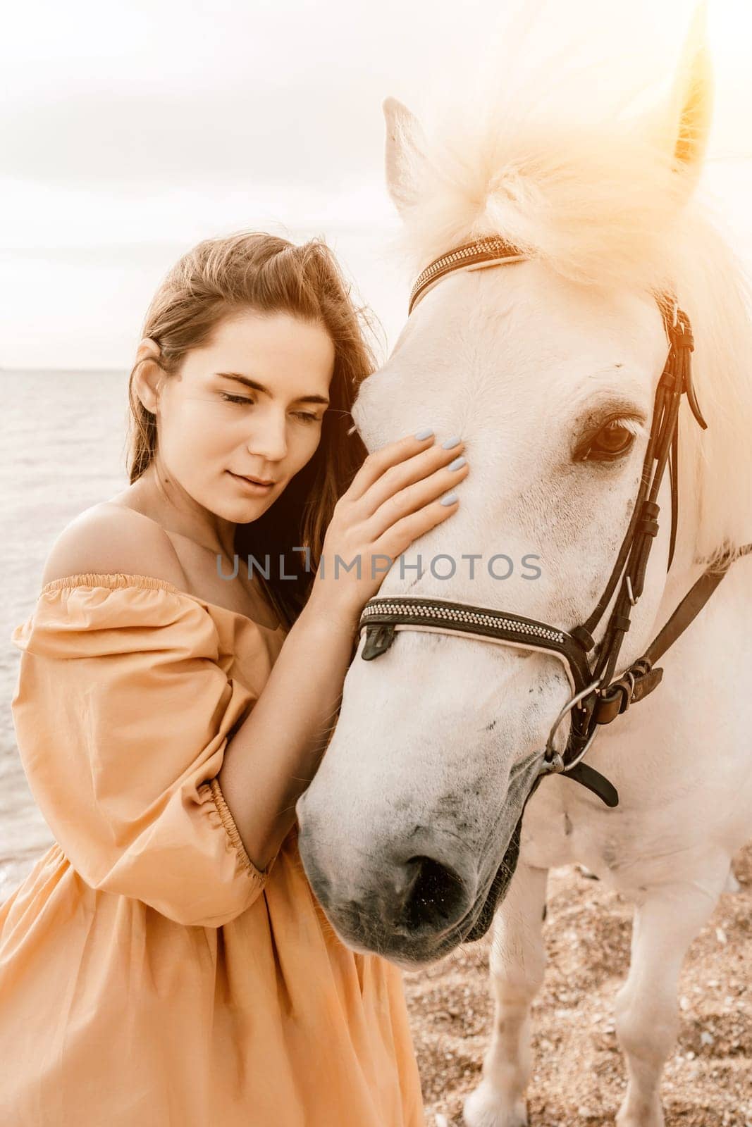 A woman in a dress stands next to a white horse on a beach, with the blue sky and sea in the background