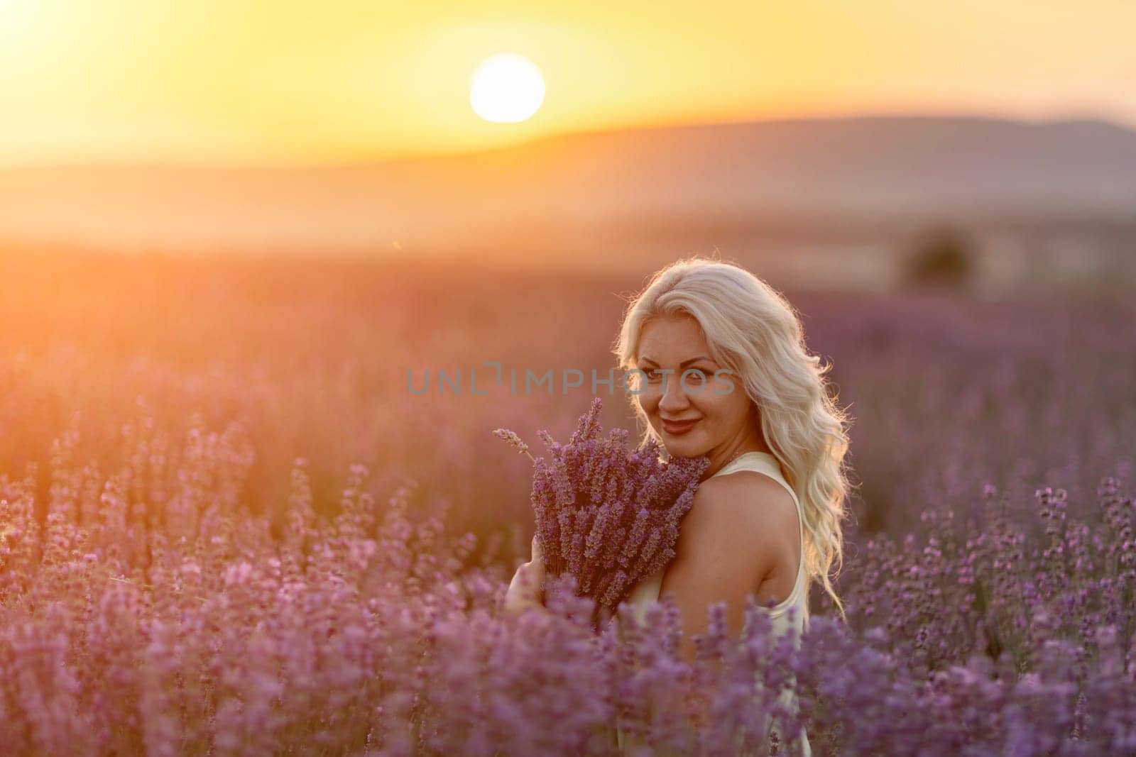 Blonde woman poses in lavender field at sunset. Happy woman in white dress holds lavender bouquet. Aromatherapy concept, lavender oil, photo session in lavender by Matiunina