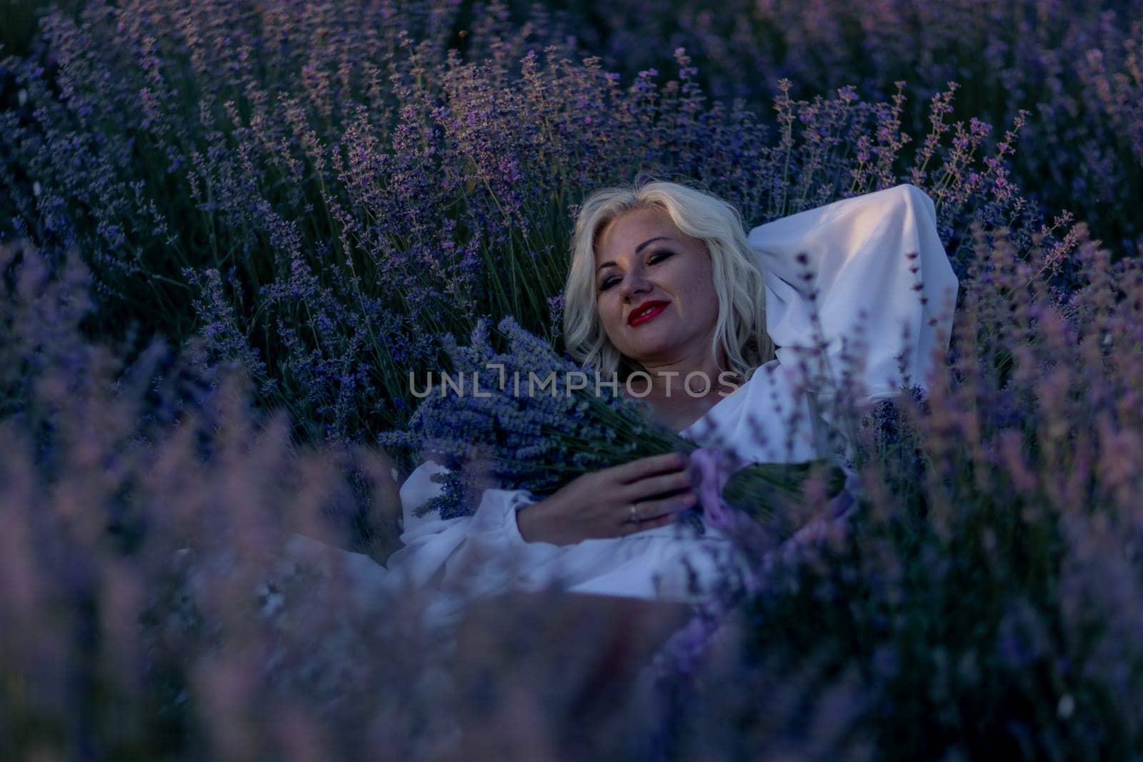 Blonde woman poses in lavender field at sunset. Happy woman in white dress holds lavender bouquet. Aromatherapy concept, lavender oil, photo session in lavender.