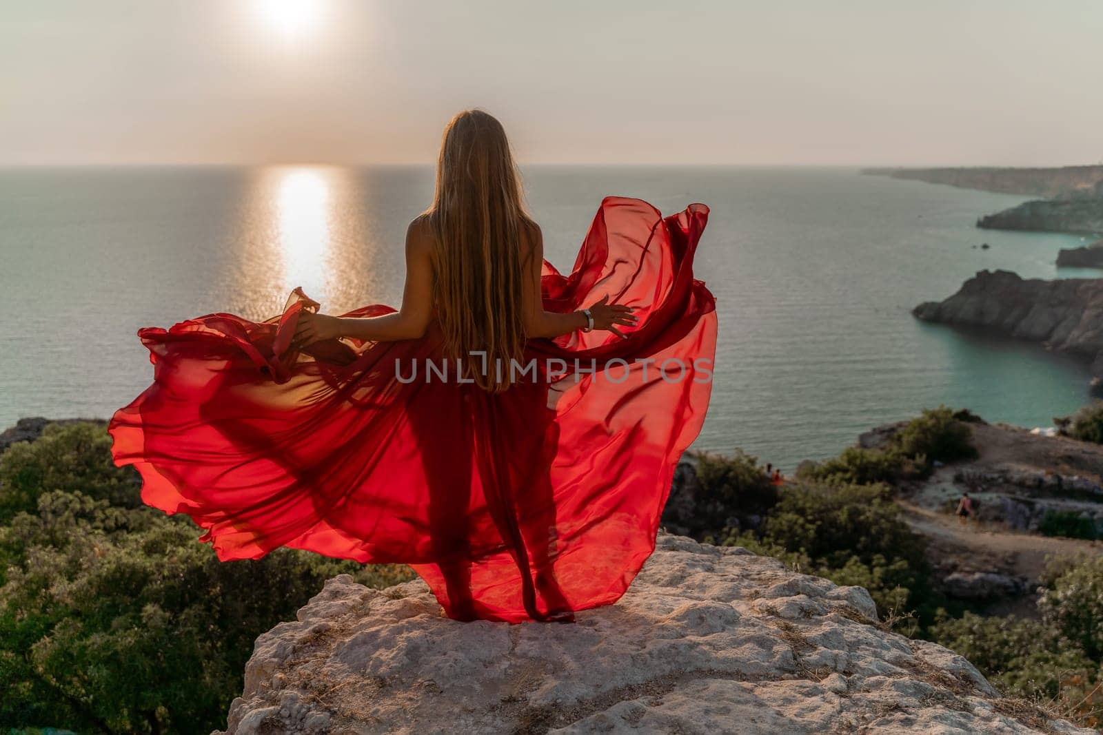 Woman sunset sea red dress, side view a happy beautiful sensual woman in a red long dress posing on a rock high above the sea on sunset