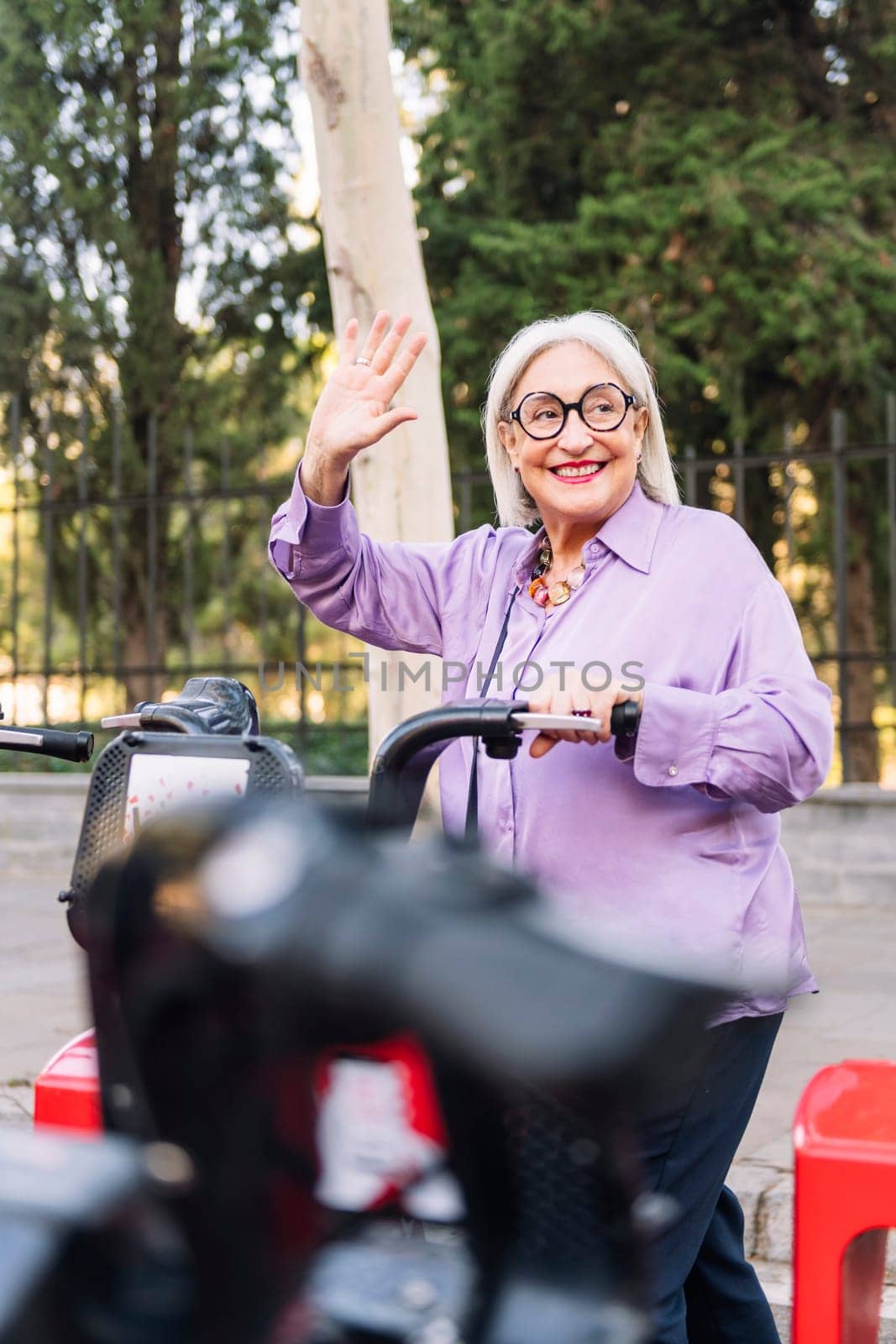 smiling senior woman greeting with hand while grabbing a rental bike from the parking row, concept of sustainable mobility and active lifestyle in elderly people