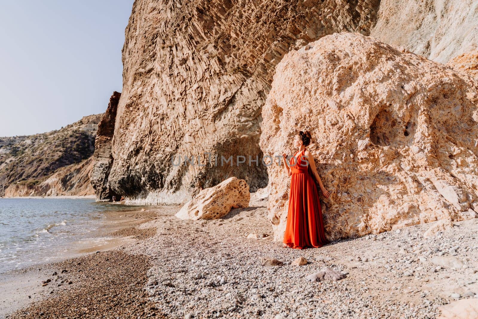 Woman red dress sea. Woman in a long red dress posing on a beach with rocks on sunny day. Girl on the nature on blue sky background