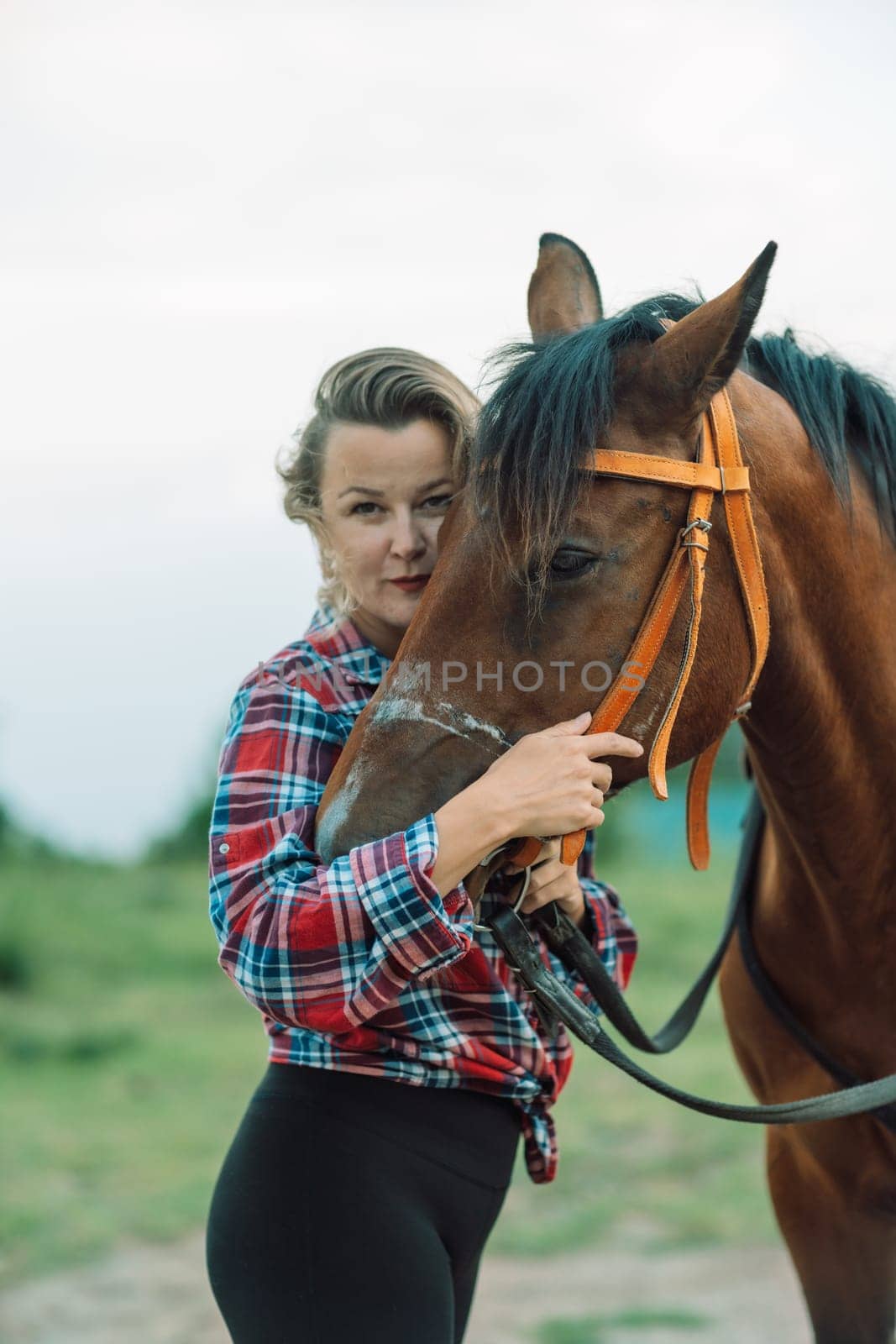 Happy blonde with horse in forest. Woman and a horse walking through the field during the day. Dressed in a plaid shirt and black leggings