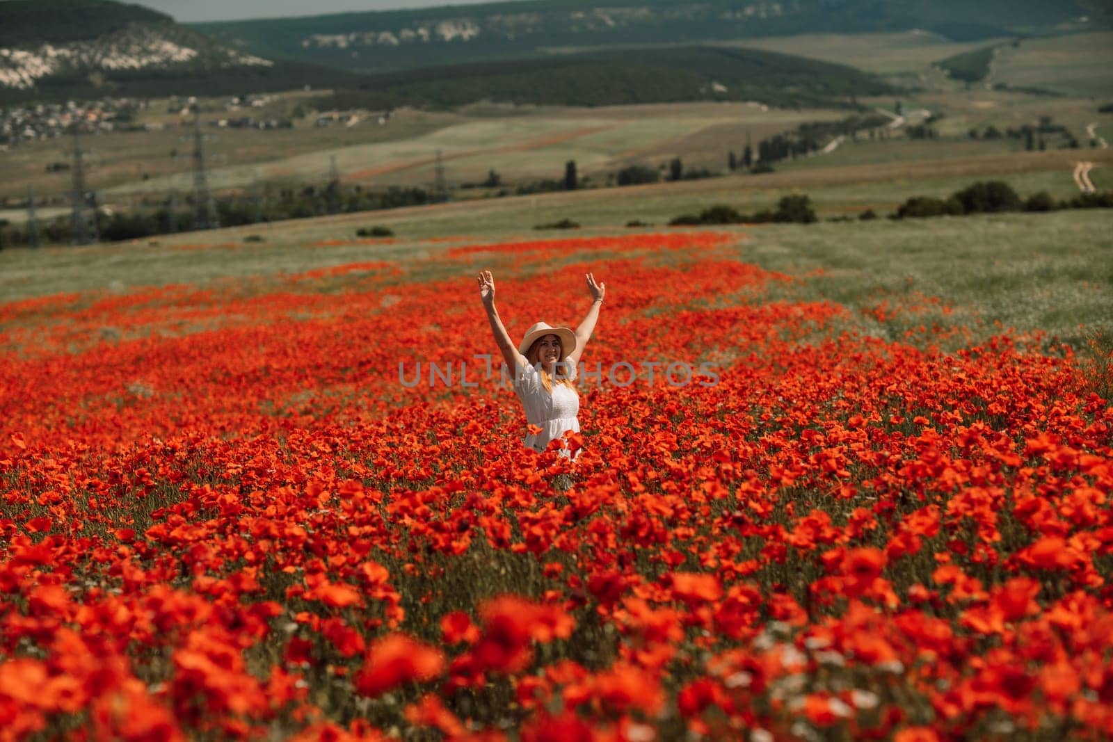 Field poppies woman. Happy woman in a white dress and hat stand through a blooming field of poppy raised her hands up. Field of blooming poppies
