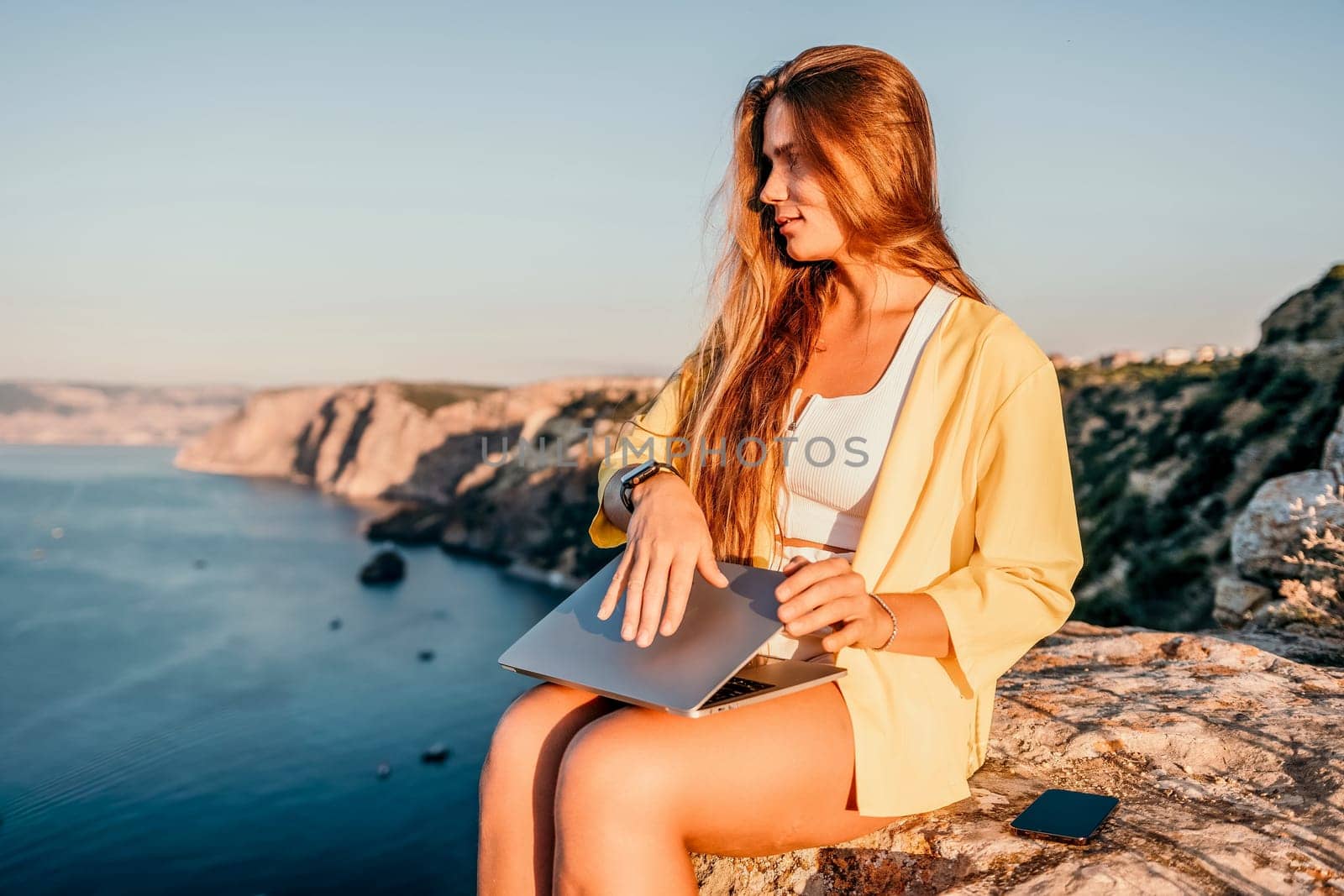 Successful business woman in yellow hat working on laptop by the sea. Pretty lady typing on computer at summer day outdoors. Freelance, travel and holidays concept.