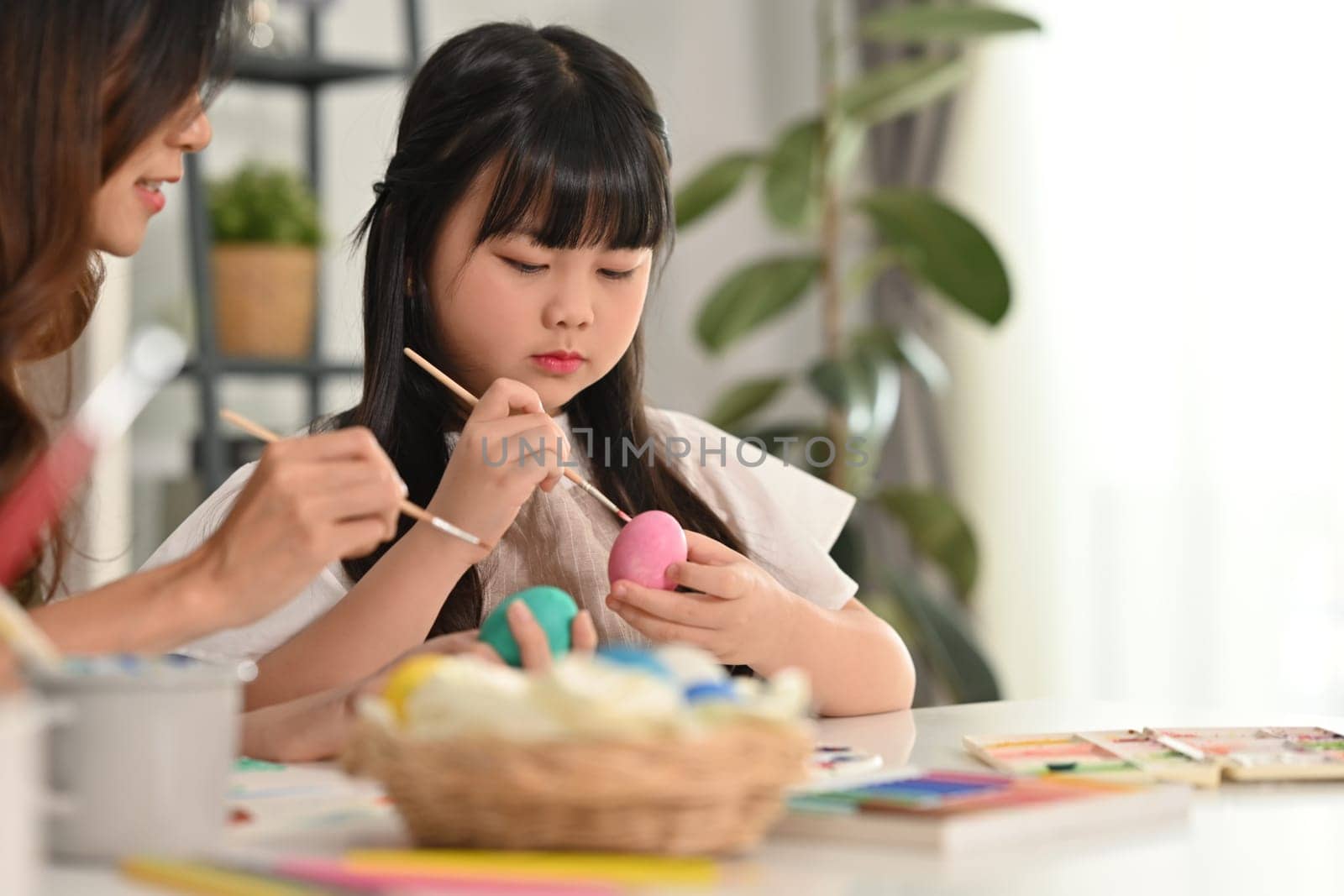 Cute little girl painting eggs with her mother, preparing decorations for easter celebration at home.