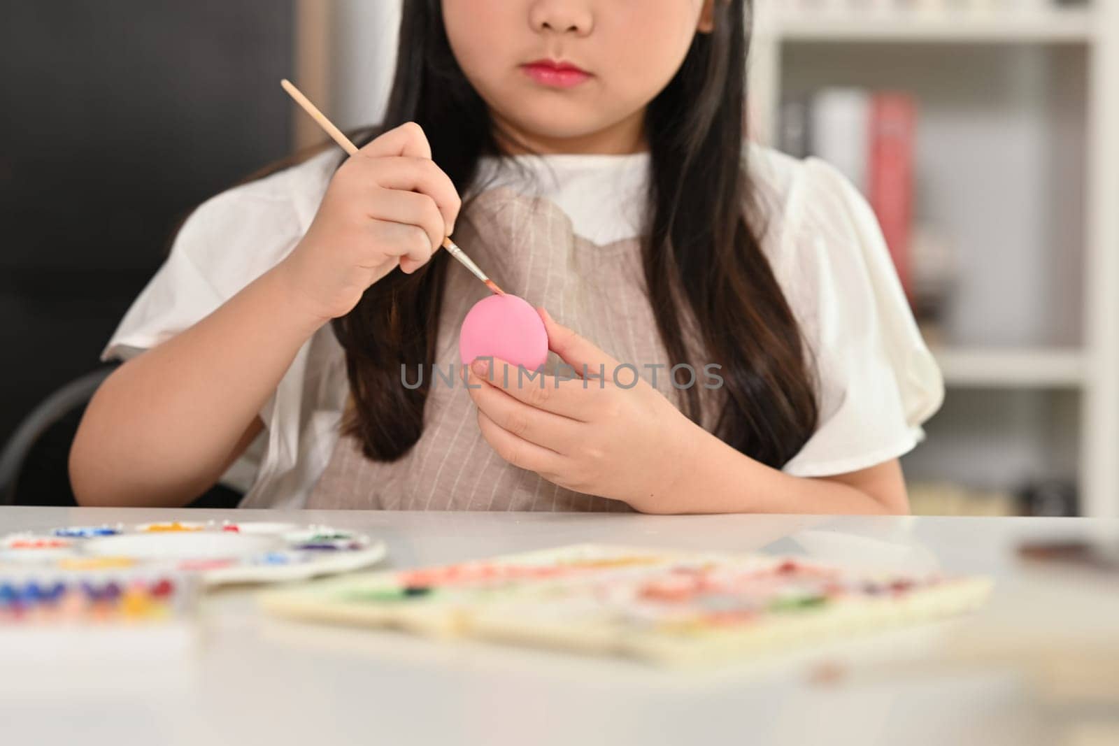 Child girl painting Easter egg with brush at table. Preparing for Easter concept. by prathanchorruangsak