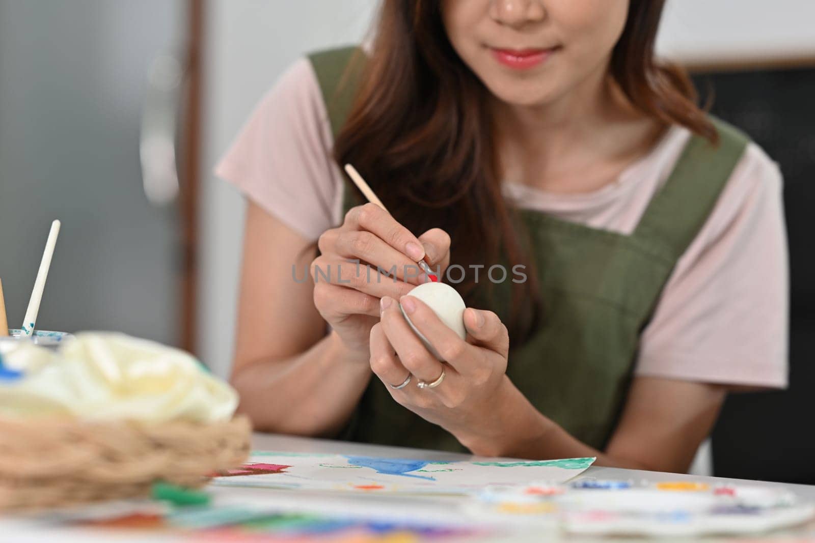 Smiling young woman in apron painting Easter egg with brush at table. Preparing for Easter concept.