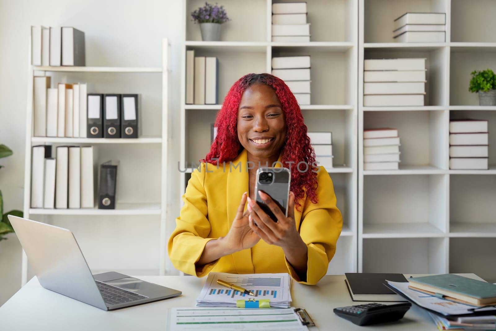 African businesswoman using smartphone phone app for playing games, shopping online, and ordering delivery while working on a laptop computer in the office.