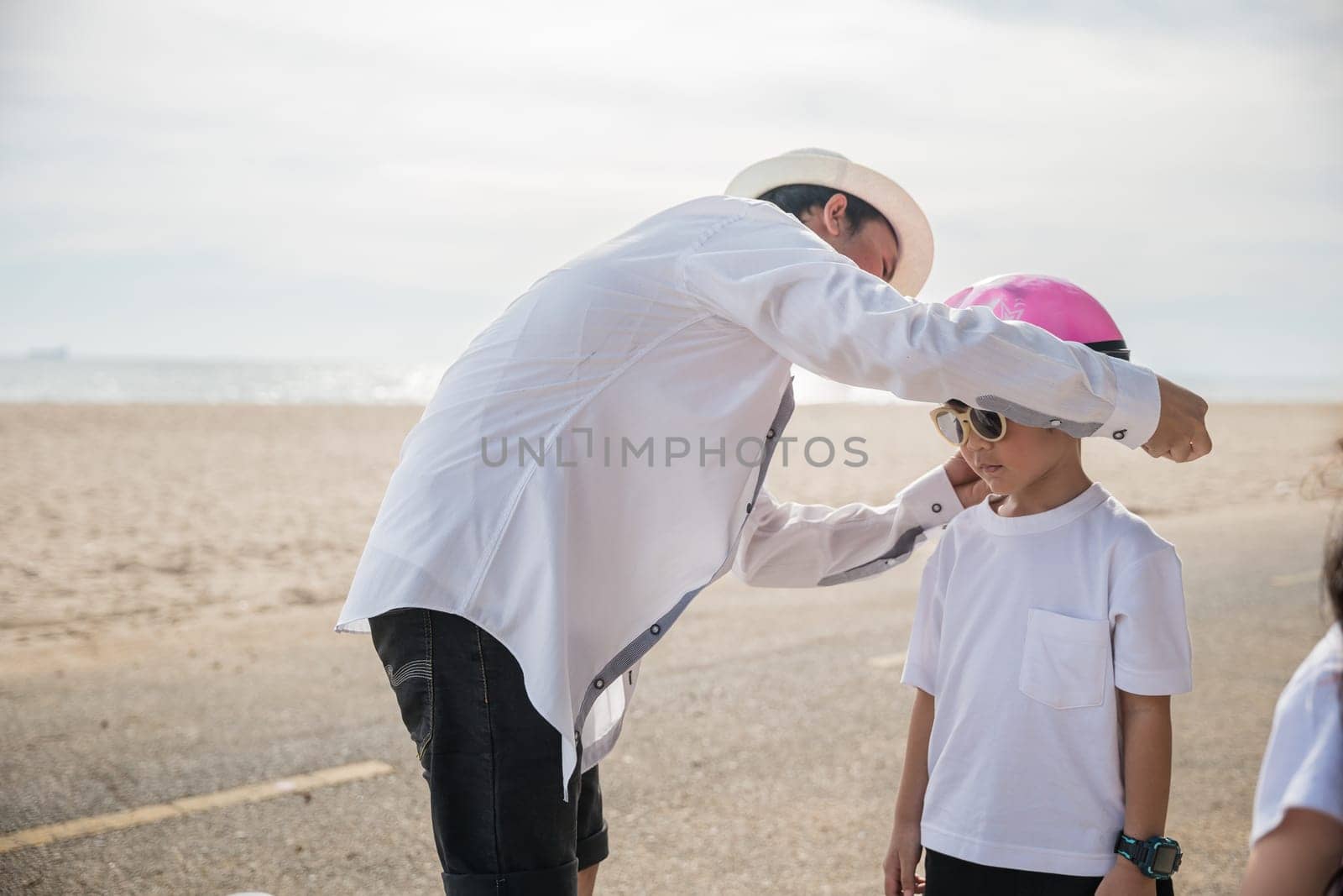 A family on vacation at the beach where the father sporting a safety helmet teaches his cheerful son the balance and joy of bicycle riding a memorable tourism day filled with family happiness.