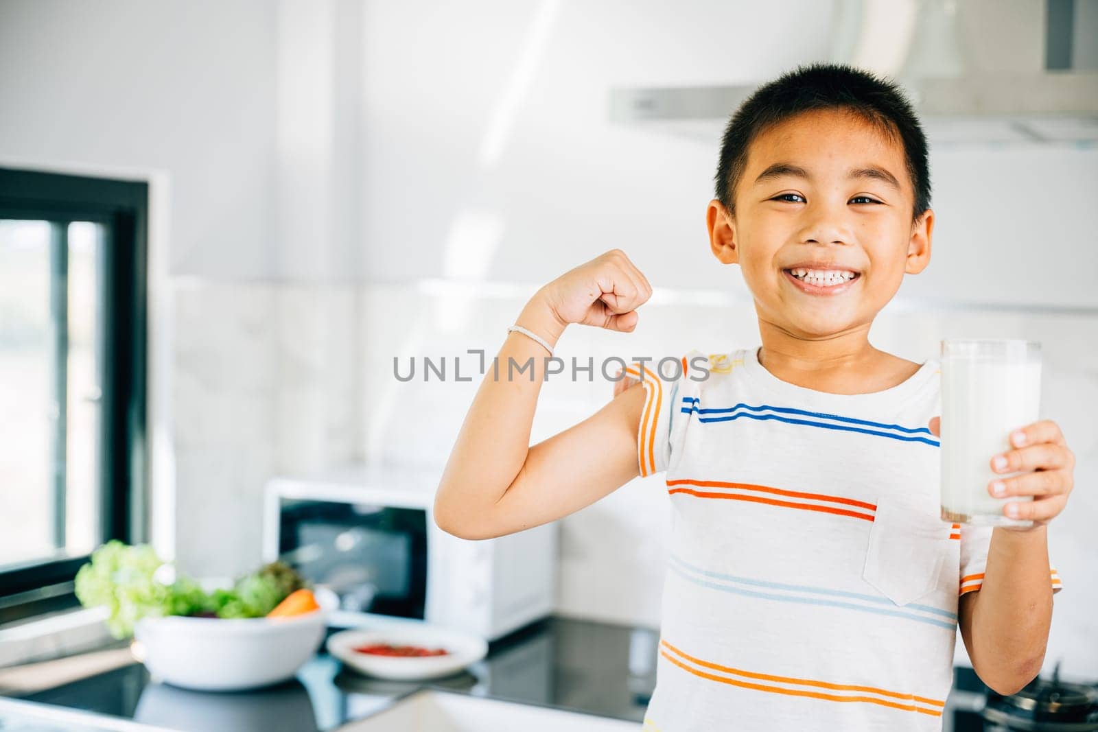 Portrait of Asian little boy in kitchen holding milk cup. Cute son enjoys drink, smiling happily. Preschool child savoring calcium-rich liquid, radiating joy at home give me. by Sorapop