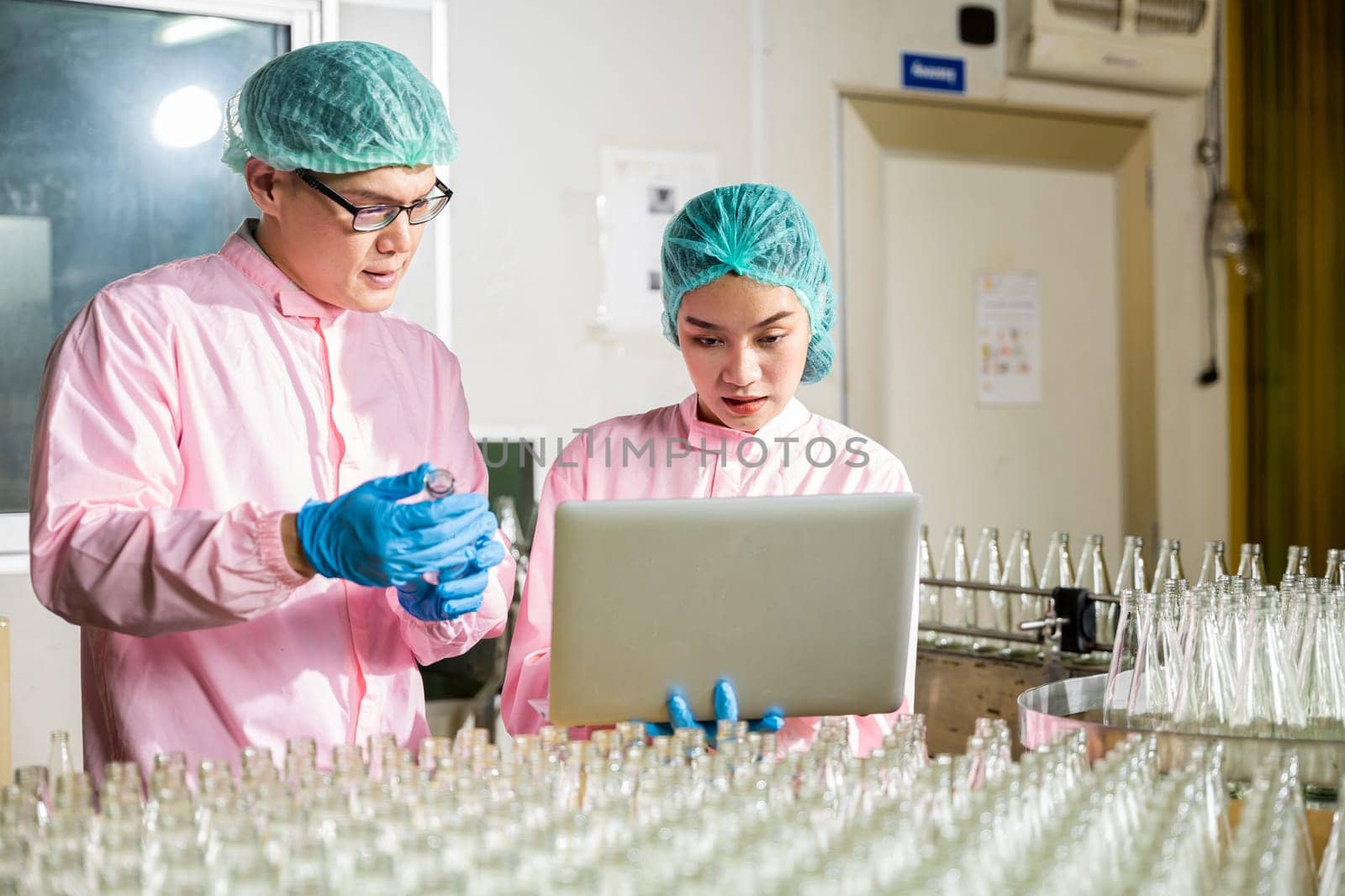 In a beverage factory's production line engineers carefully inspect product bottles on conveyor belt. Inspection and quality control conducted by professionals using a laptop for meticulous checks.