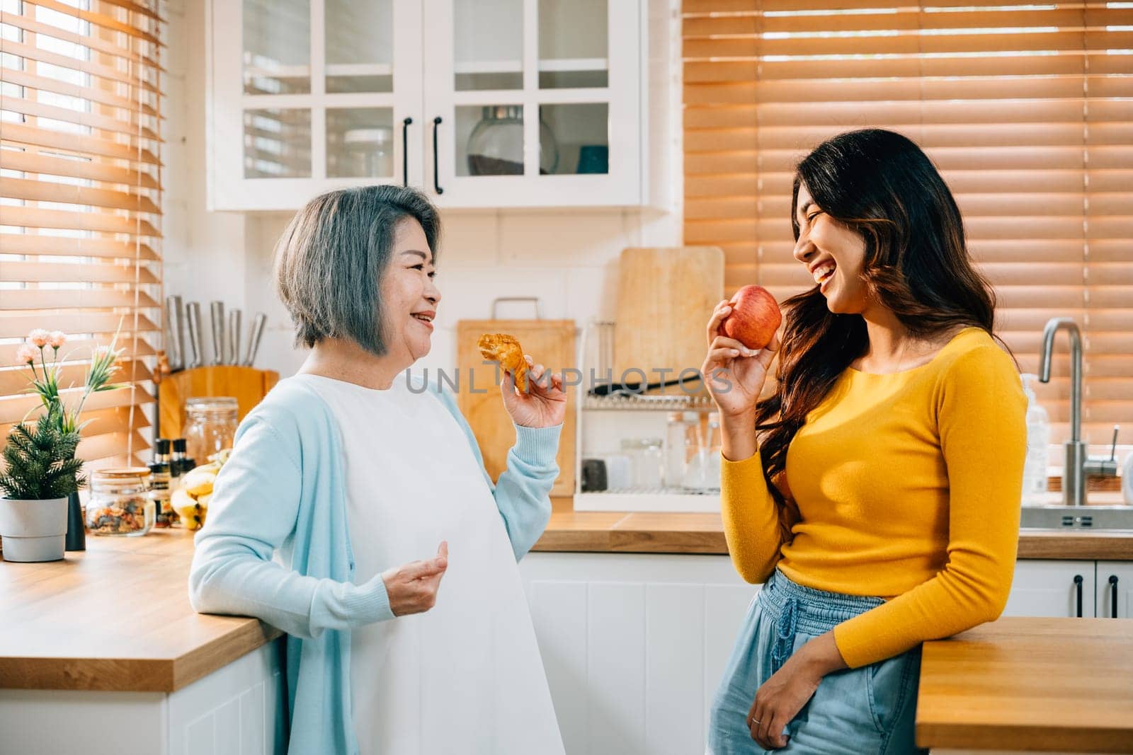 An elderly mother and her adult daughter, a young woman in an apron, bond over food in the kitchen. Their cheerful smiles highlight the happiness and fun of family togetherness at home. by Sorapop