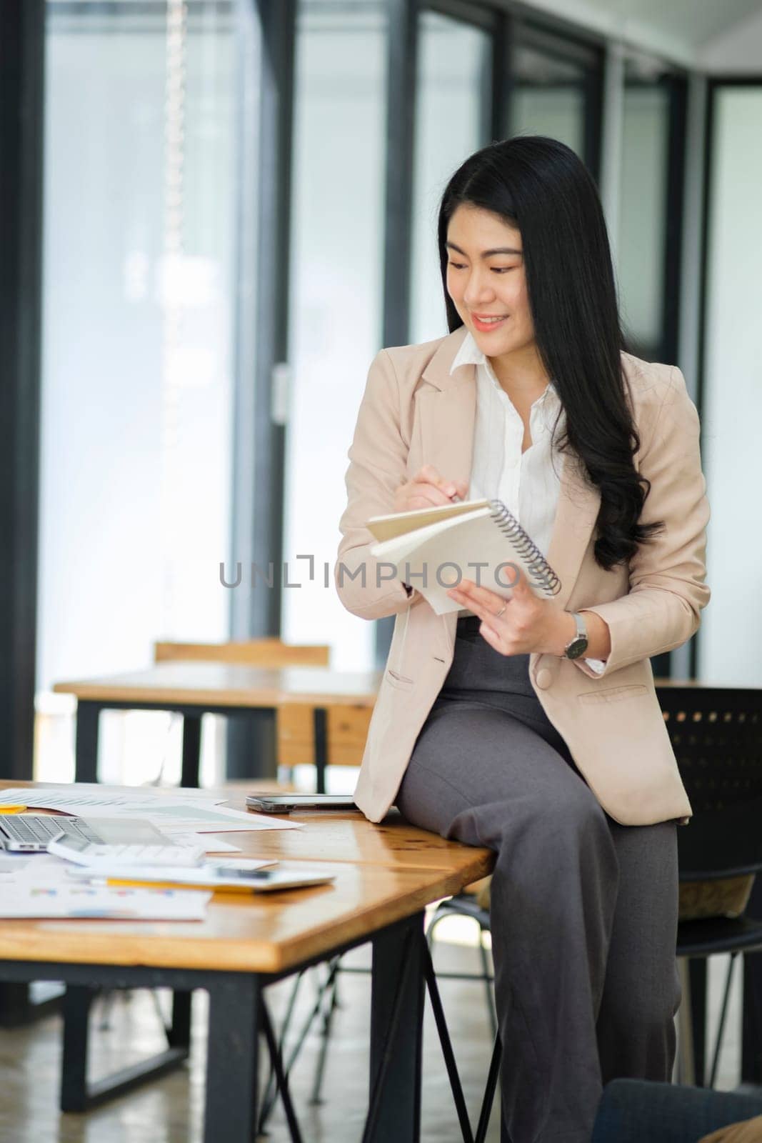 A focused businesswoman working on a laptop while holding a notebook in a bright office environment..