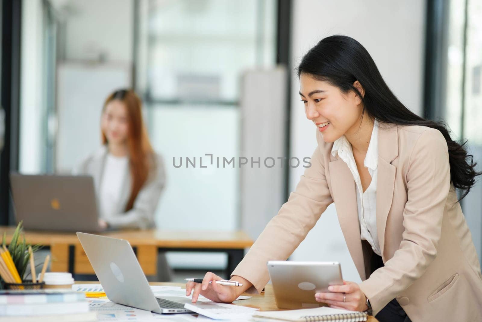 A focused businesswoman working on a laptop while holding a notebook in a bright office environment..