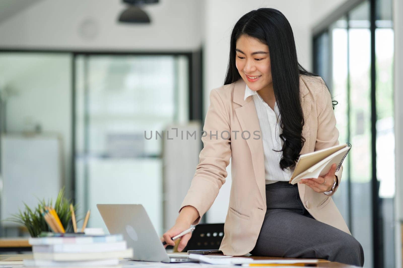 A focused businesswoman working on a laptop while holding a notebook in a bright office environment..