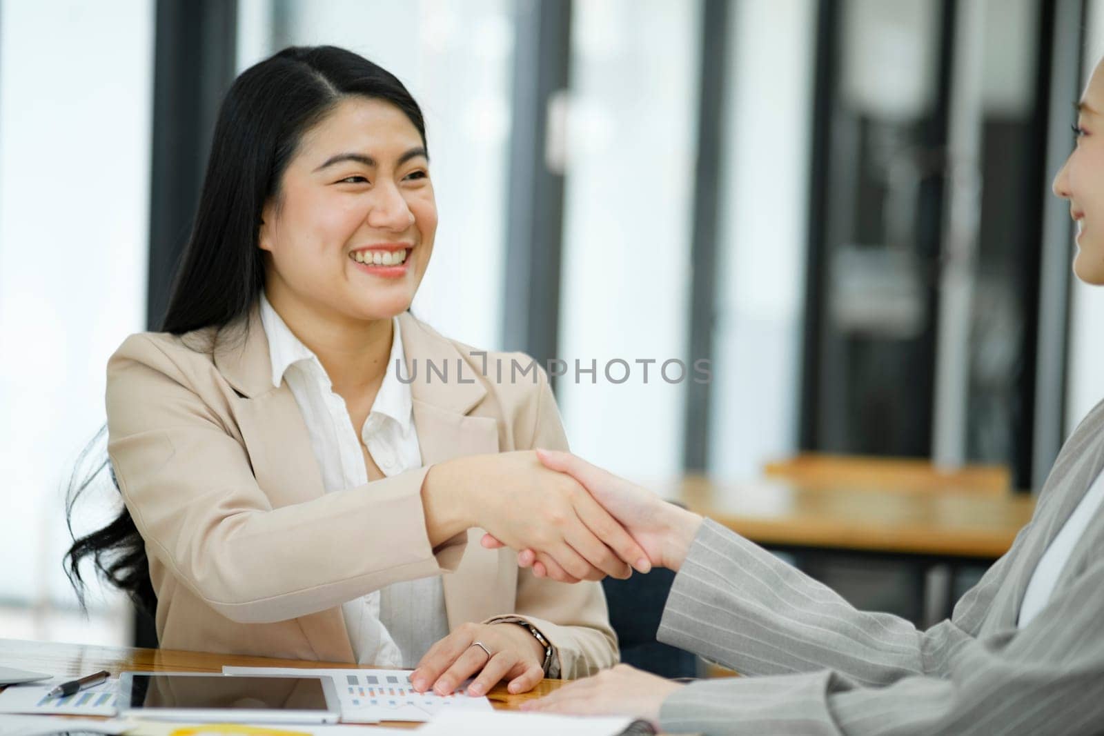 Two smiling businesswomen shaking hands over a desk in the office, symbolizing a successful agreement or partnership.