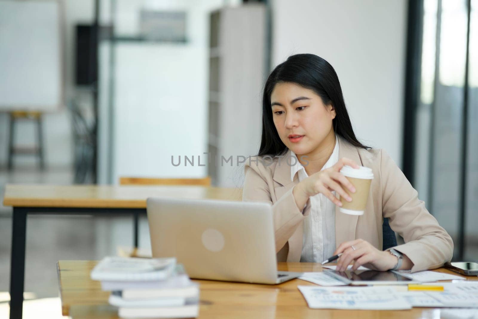 A focused businesswoman working on a laptop while holding a notebook in a bright office environment..