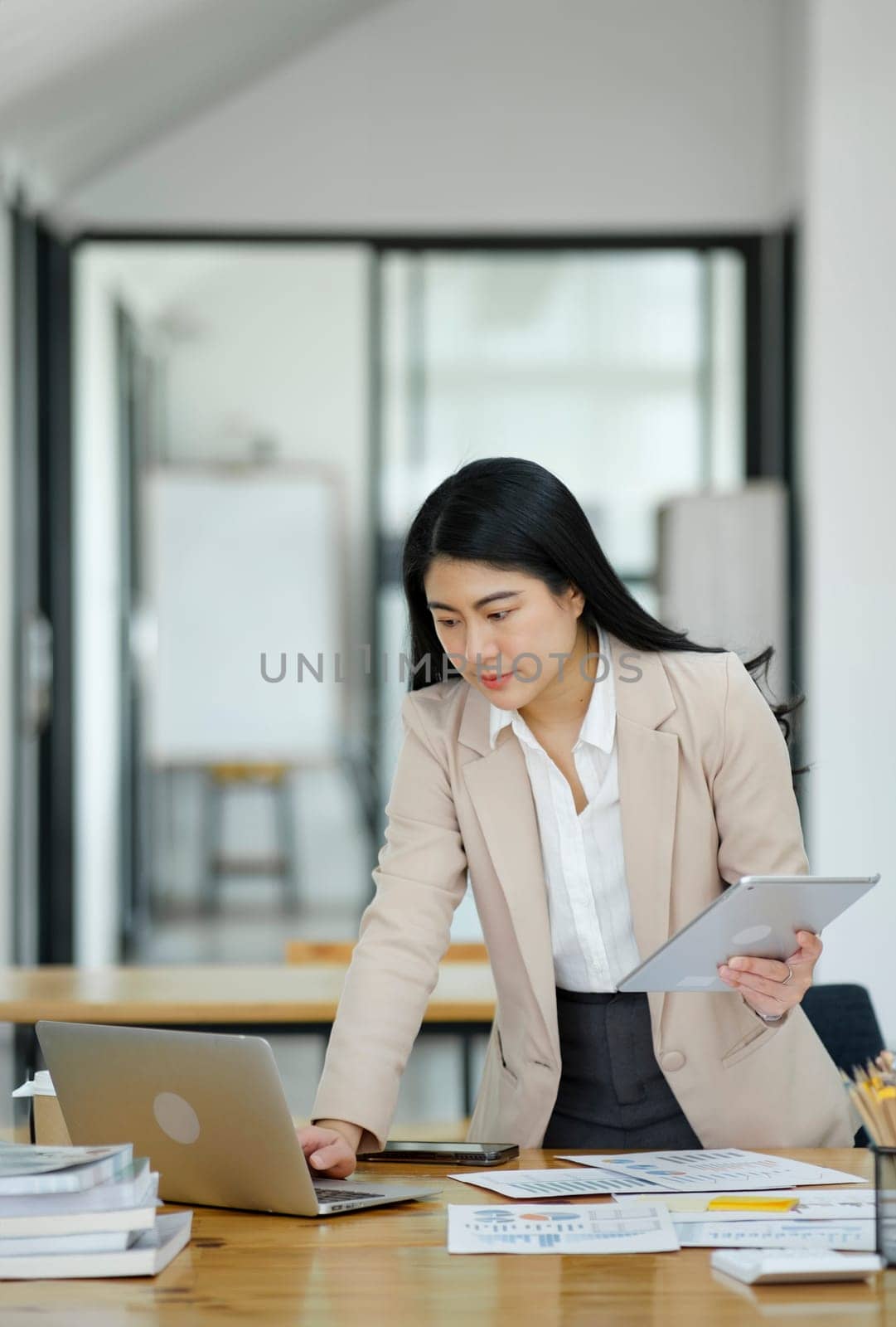 A focused businesswoman working on a laptop while holding a notebook in a bright office environment..