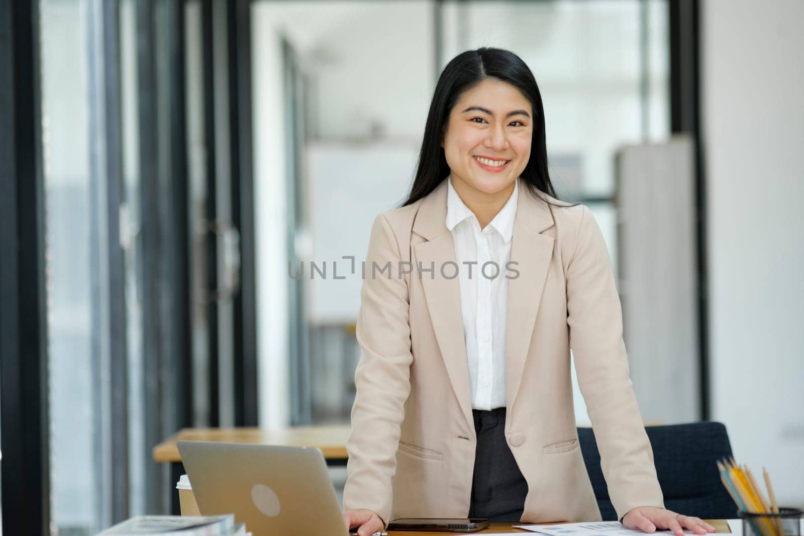 A professional and confident businesswoman smiling at the camera, standing by her desk with a laptop in a modern office.