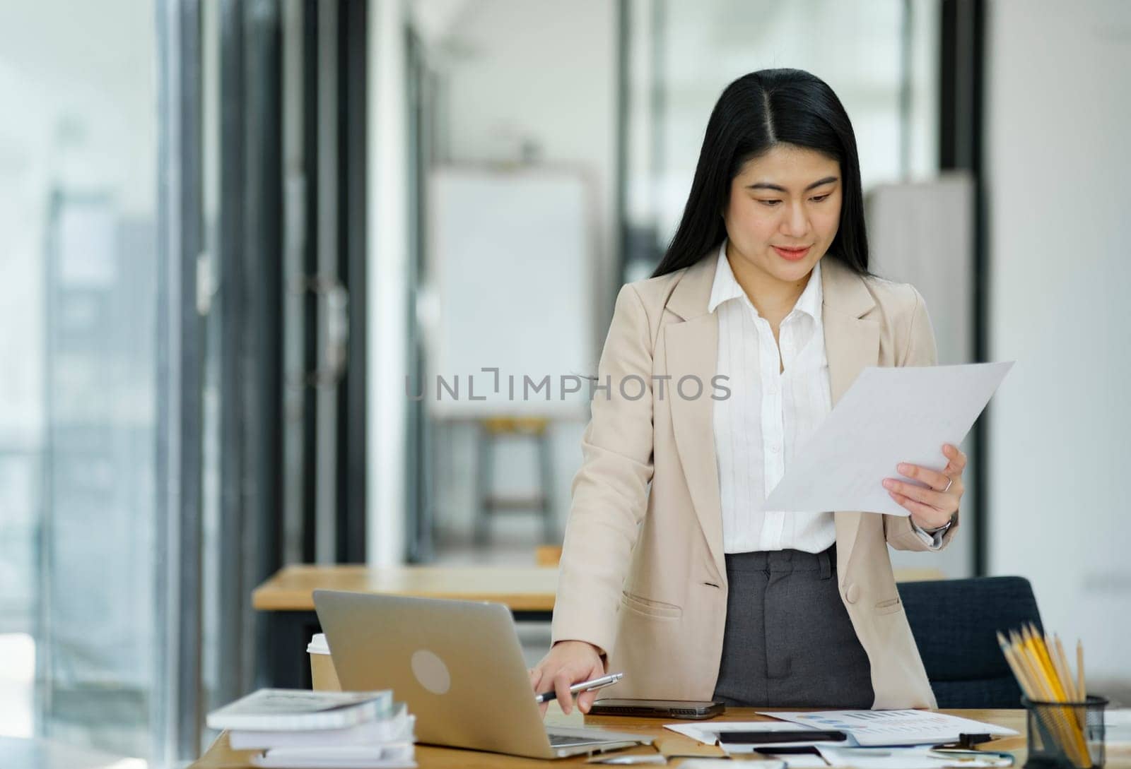 A focused businesswoman working on a laptop while holding a notebook in a bright office environment..