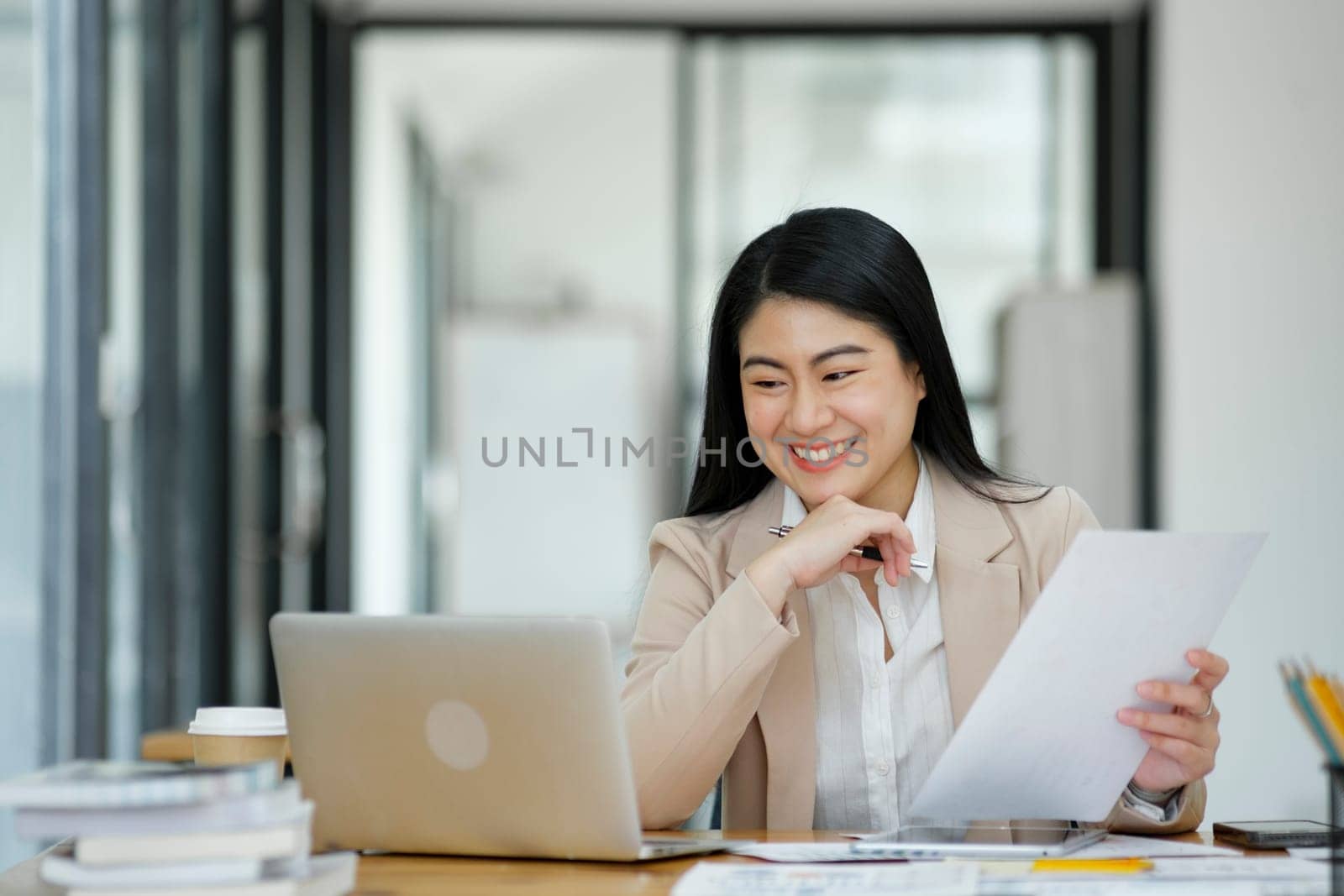 A focused businesswoman working on a laptop while holding a notebook in a bright office environment..