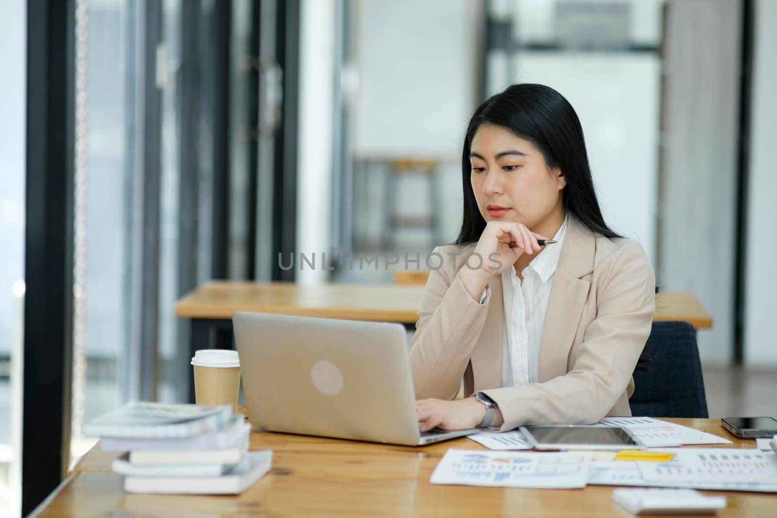 A focused businesswoman working on a laptop while holding a notebook in a bright office environment..